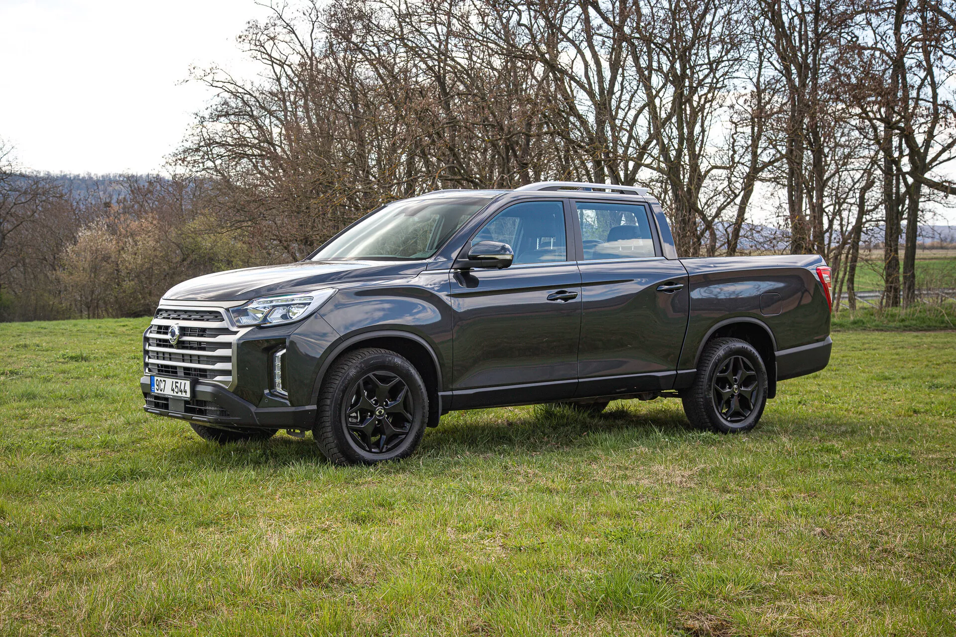 Boční pohled na tmavě šedý pickup SsangYong Musso Grand 2022 na travnatém poli, ukazuje jeho slitinová kola, LED světlomety a prostornou nákladní ložnou plochu