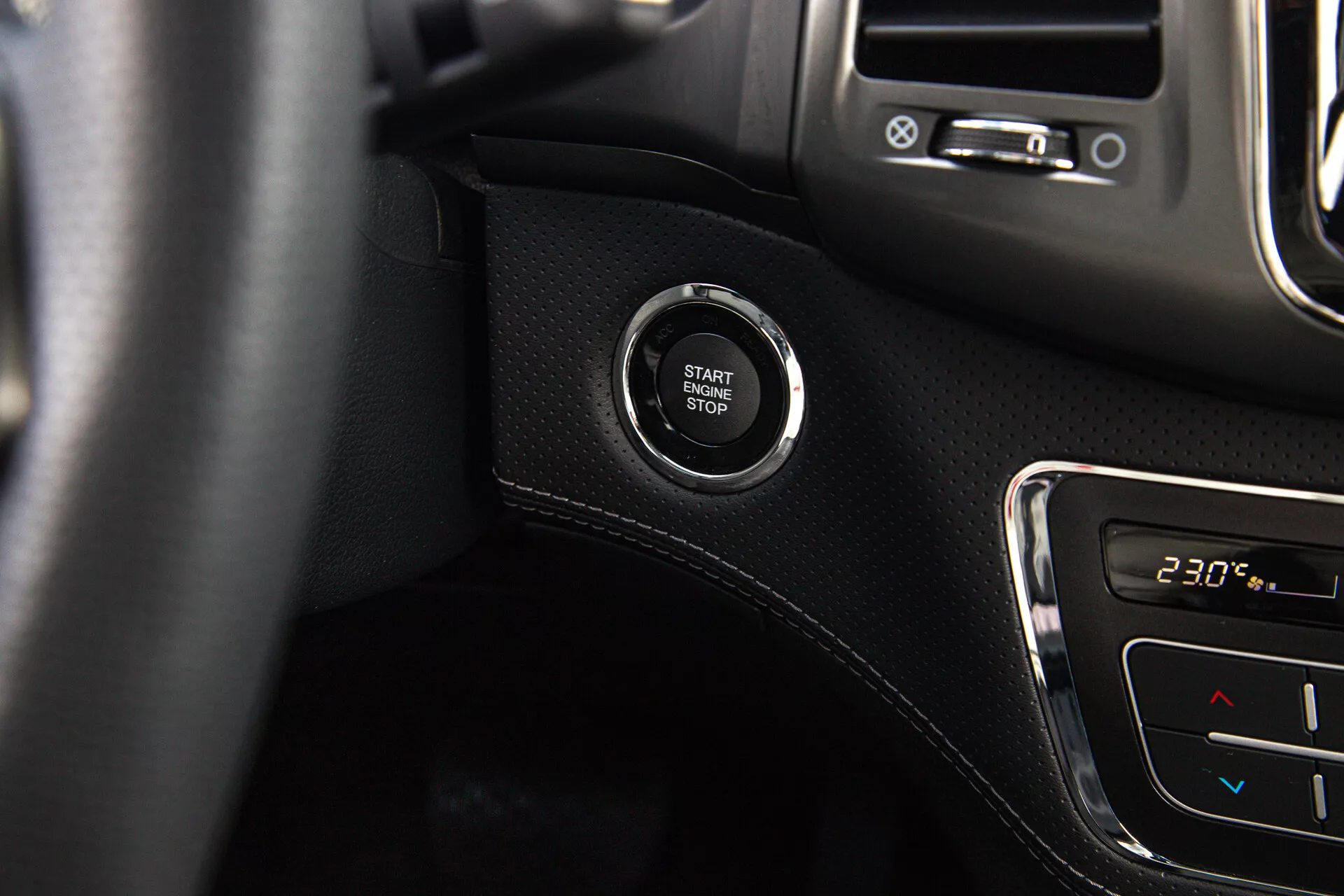Detailní pohled na tlačítko Start/Stop motoru v SsangYong Musso 2022, zdůrazňující systém bezklíčového zapalování na palubní desce s černým povrchem na pozadí a detaily klimatizace