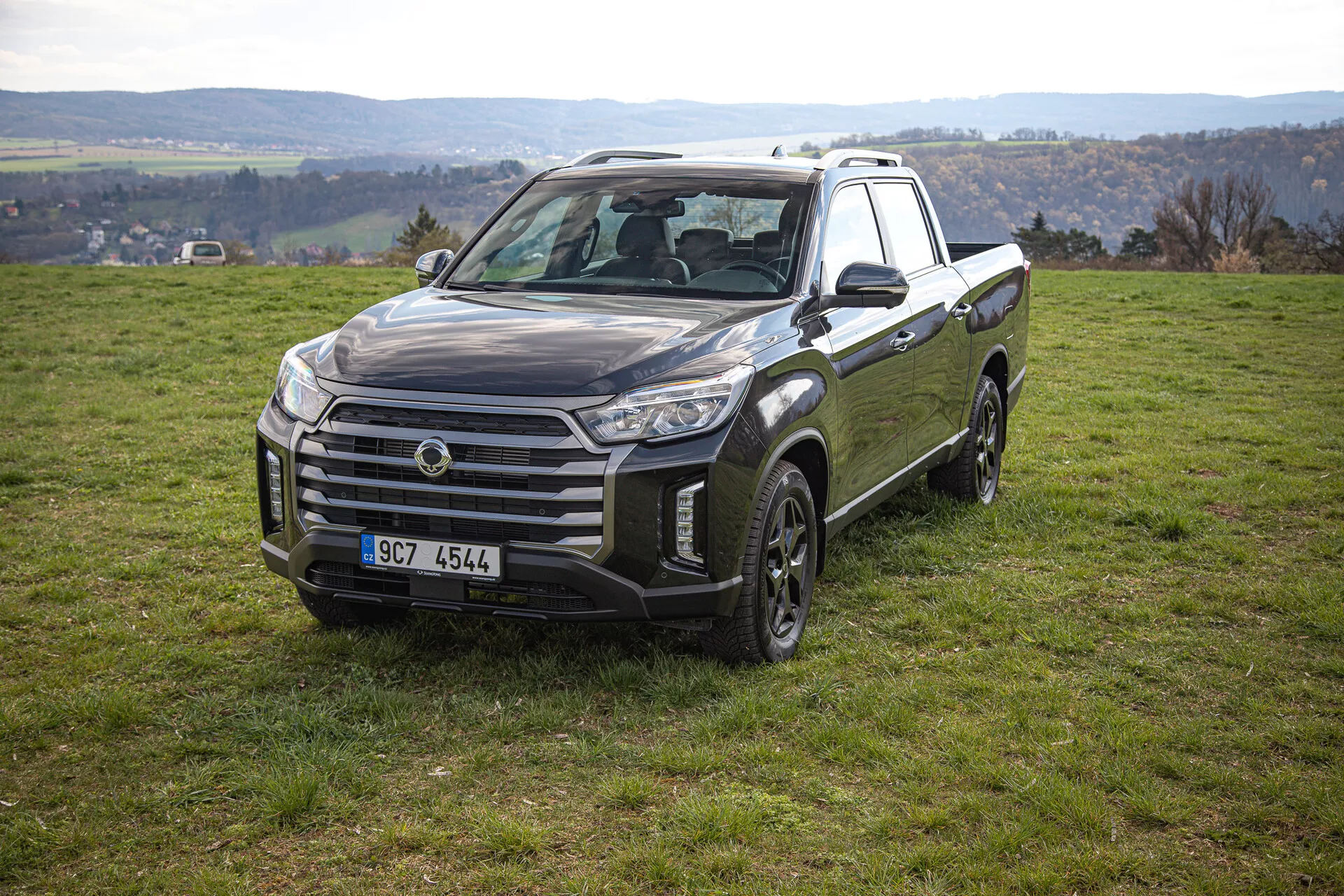 Stacionárně zobrazený pickup SsangYong Musso Grand 2022 na mírném svahu s trávou s výhledem na přední a boční stranu včetně velké mřížky, úhlových světlometových sestav a designu kol vhodného pro jízdu v terénu, v pozadí s venkovskou krajinou.