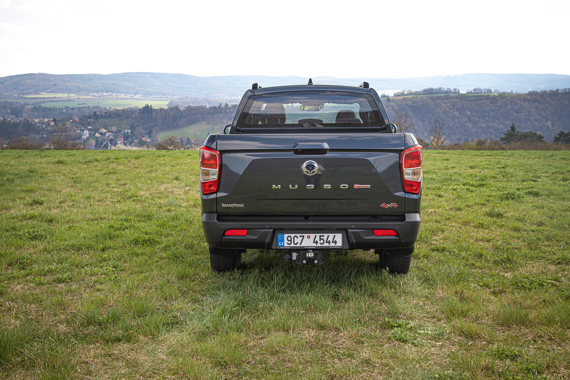 Zadní pohled na tmavý pickup SsangYong Musso Grand 2022 na nerovném trávníku s pozadím venkovské krajiny, předvádějící značení na zadních vratkách, vertikální zadní světla, zadní senzory a tažné zařízení během dne