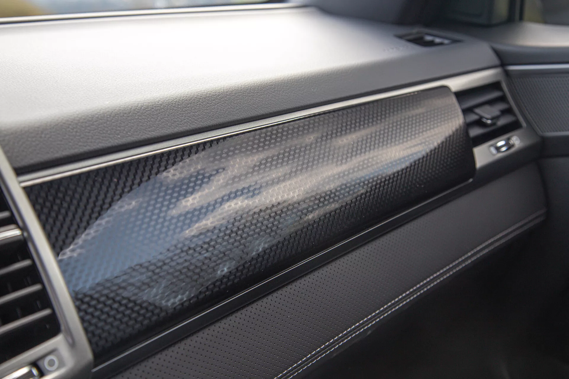 Detailní zobrazení palubní desky SsangYong Musso 2022 s luxusním karbonovým obložením a materiály vysoké kvality, zaměřené na stranu spolujezdce s náznaky schránky na rukavice, větrací mřížky a ovládání teploty, ilustrující důraz vozidla na moderní design interiéru a zpracování.