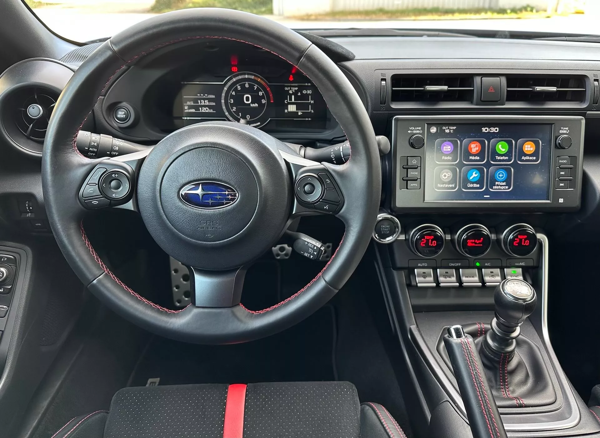 Interiér Subaru BRZ 2023 z perspektivy řidiče obsahující sportovní kožený volant s multifunkčními ovládáními a červeným prošíváním, přístrojovou desku s analogovými ciferníky a digitálním displejem, středovou konzolu s velkým dotykovým infotainment systémem a duální klimatizací, a manuální řadící páku s koženým potahem a červenými akcenty, na pozadí černého interiéru se sportovními červenými akcenty, zdůrazňující pokročilé funkce a technologie vozidla.