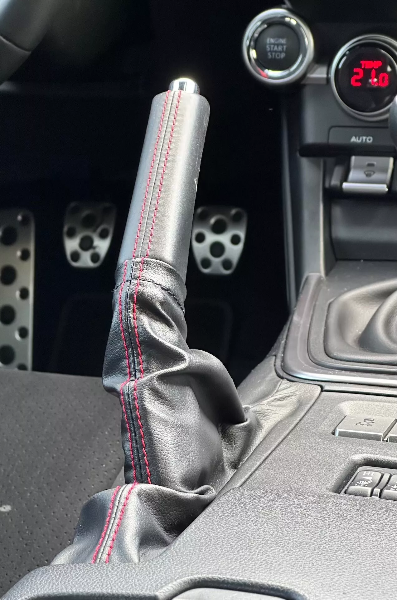 Detailní pohled na středovou konzolu Subaru BRZ 2023, zdůrazňující kovovou řadicí páku s červeným kontrastním prošíváním na koženém obalu, digitální údaj zobrazující '21', sportovní hliníkové pedály s vyvrtanými otvory a technologické funkce, jako jsou tlačítka pro elektronickou stabilizaci.