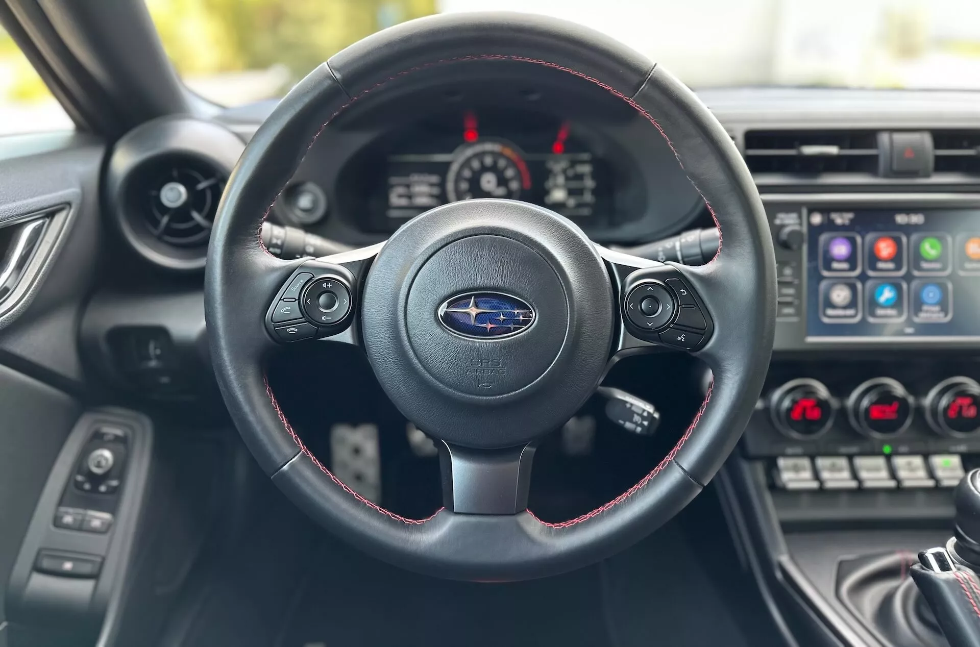 Interiér Subaru BRZ 2023 z pohledu řidiče zaměřený na tříramenný volant s červeným prošíváním, logo Subaru a palubní desku s přístrojovým panelem a středovou konzolou
