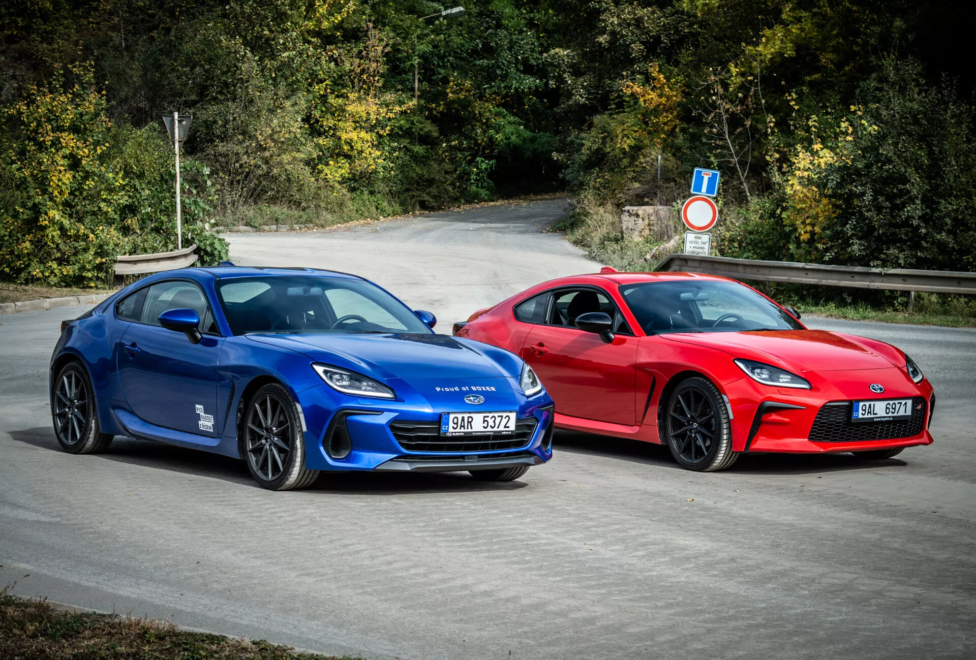 Sportovní kupé Subaru BRZ 2023 v modré a červené s aerodynamickým designem a výkonnostními prvky, zaparkované venku obklopené přírodní zelení
