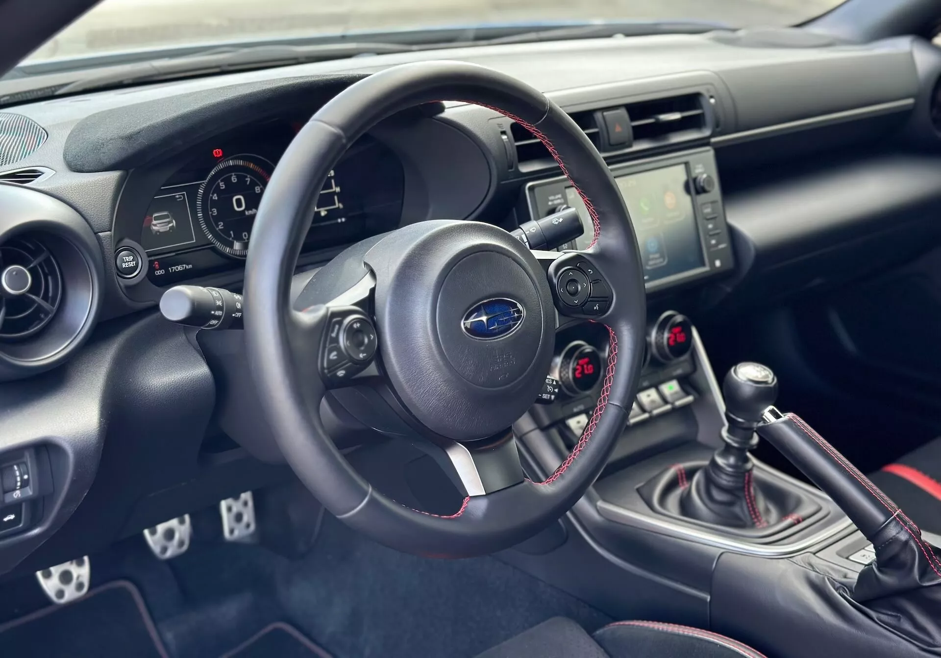 Interiér Subaru BRZ 2023 s důrazem na palubní desku a kokpit řidiče s detailním pohledem na volant, přístrojovou desku, středovou konzolu s ovládáním audia a klimatizace a manuální řadicí páku se sportovním červeným prošíváním