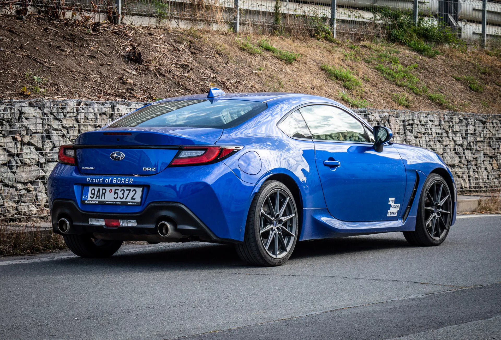 Sportovní kupé Subaru BRZ 2023 v metalické modré barvě zaparkované na sklonu, zdůrazňující zadní tříčtvrteční pohled vozidla, boční profil, zakřivený zadek, sportovní slitinová kola, dvojité výfukové hroty, malý spoiler na zavazadlovém víku a odznak motoru 'Proud of Boxer'.