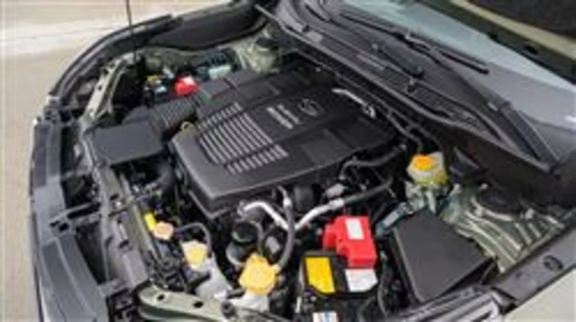 Detailní pohled na motorový prostor Subaru Forester 2015 s baterií, nádržkami na kapaliny, krabicí vzduchového filtru, různými potrubími a hadicemi, mechanickými díly a krytem motoru