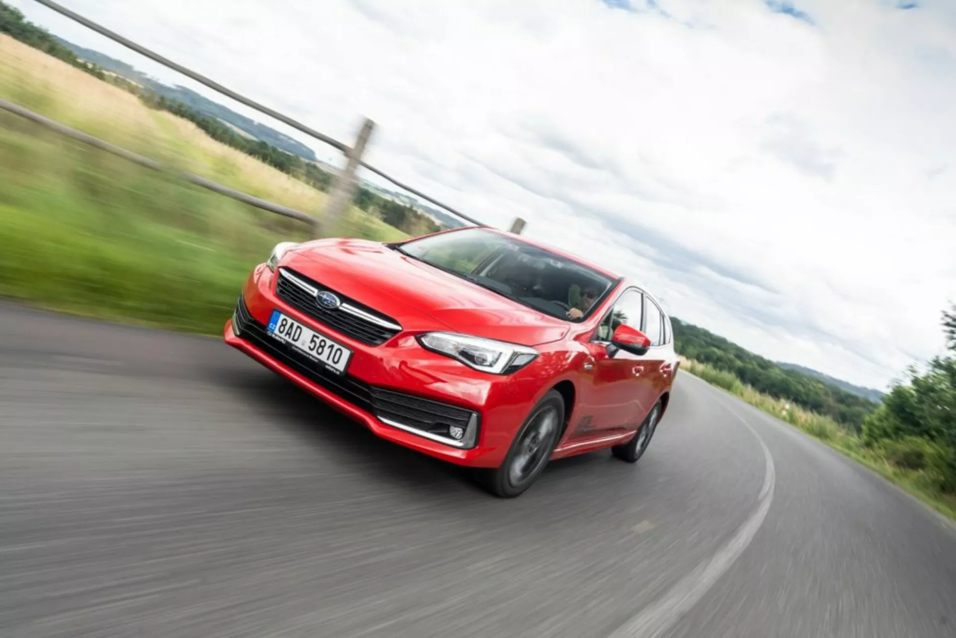 Dynamický obrázek s rozmazáním pohybu červeného Subaru Impreza z roku 2018 v akci, pohled zepředu ukazující světlomet, mřížku a slitinová kola během otáčení vlevo na venkovské silnici