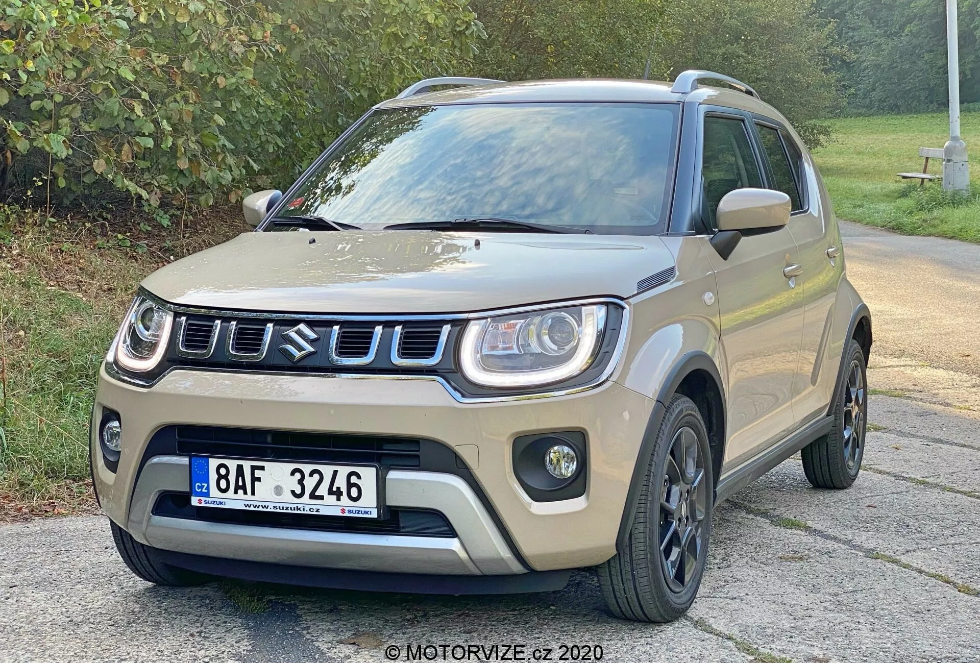 Kompaktní SUV Suzuki Ignis 1.2 Dualjet Hybrid Club z roku 2020 zobrazené ze tříčtvrtečního předního úhlu, ukazující jeho světle béžové nebo pískové tělo s černými akcenty na blatnících, dolním bočním těle a střeše. Přední mřížka má obdélníkový vzor se stříbrným nebo chromovým okrajem. Přední nárazník má výrazné kryty na mlhovky a dynamickou kapotu s mírnými konturami. Černá vícepaprsková litá kola a černá boční zrcátka dodávají automobilu robustní vzhled. Vozidlo je zaparkováno na asfaltovém povrchu s zeleným listím v pozadí pod měkkým osvětlením.