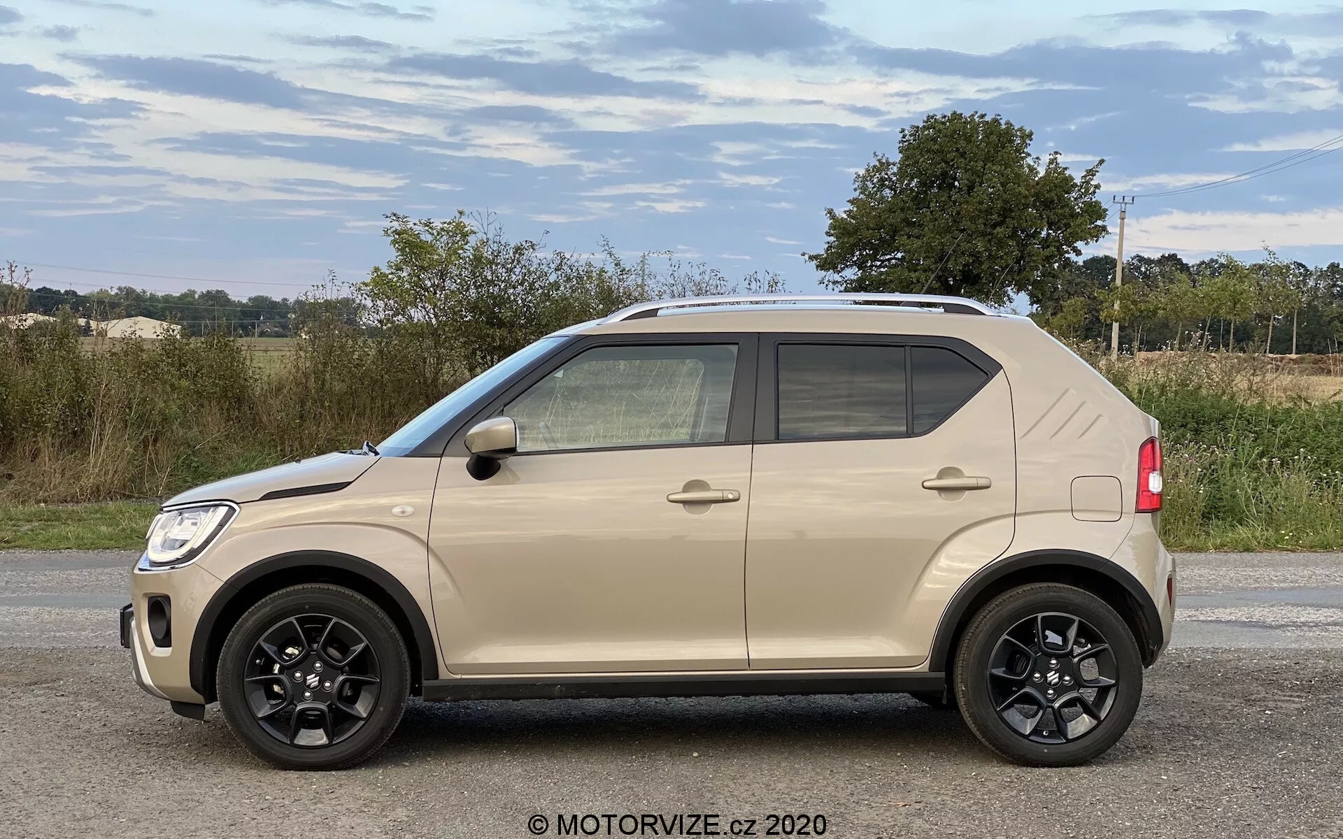 Boční profil kompaktního SUV Suzuki Ignis 1.2 Dualjet Hybrid Club z roku 2020 s viditelnými čtyřmi dveřmi, černými pětiramennými litými koly, dvoubarevným béžovo-černým designem, tónovanými okny a možnými zrcátky s integrovanými směrovými světly na venkovském pozadí.