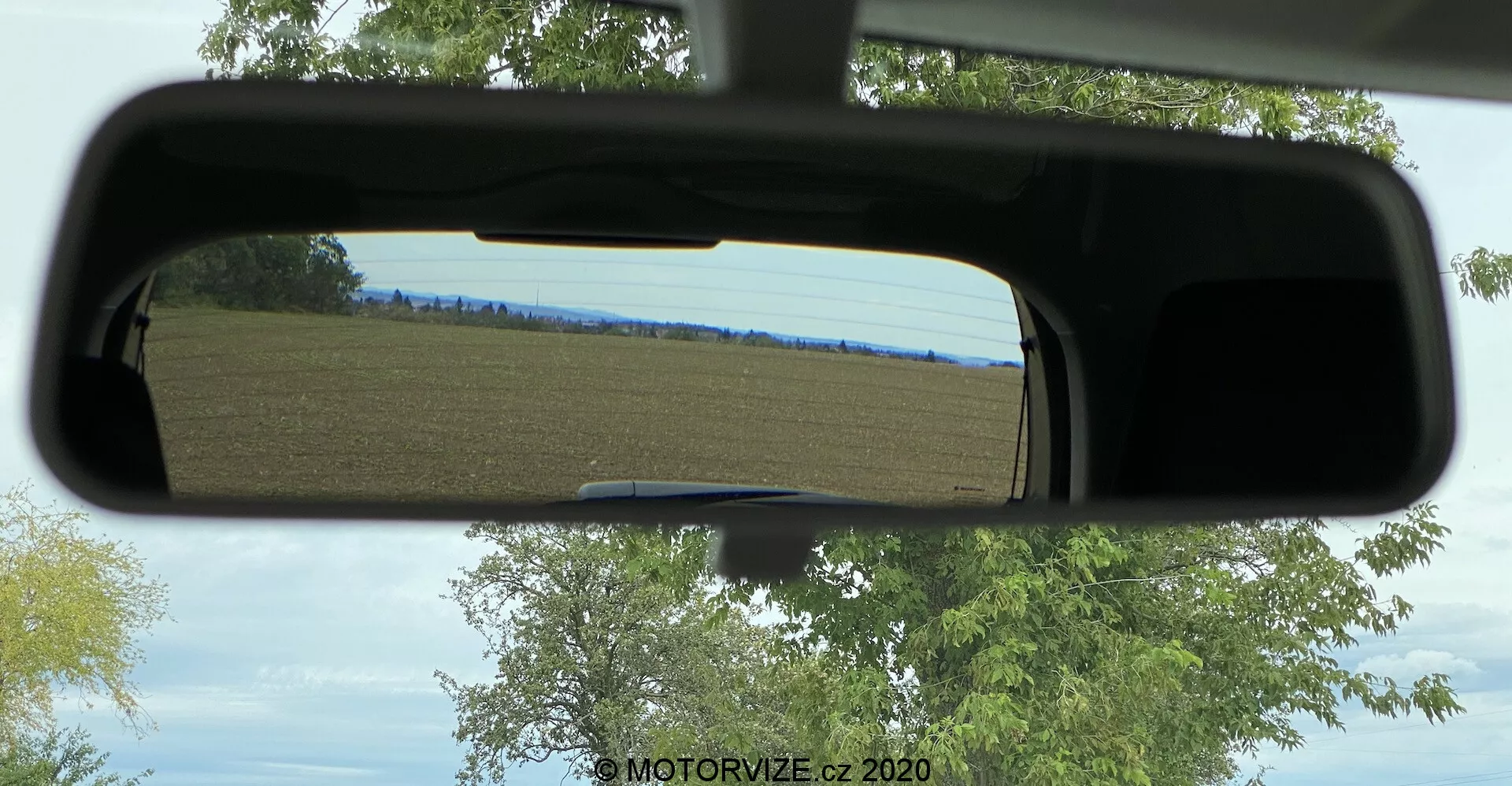 Pohled skrz standardní zpětné zrcátko zobrazující jasný den s venkovskou krajinou sestávající převážně z pole a stromů. Zrcátko je orámováno vnitřní střechou vozidla a zřejmě vrchem čelního skla, což dává pocit, jako by bylo viděno z místa řidiče nebo spolujezdce.