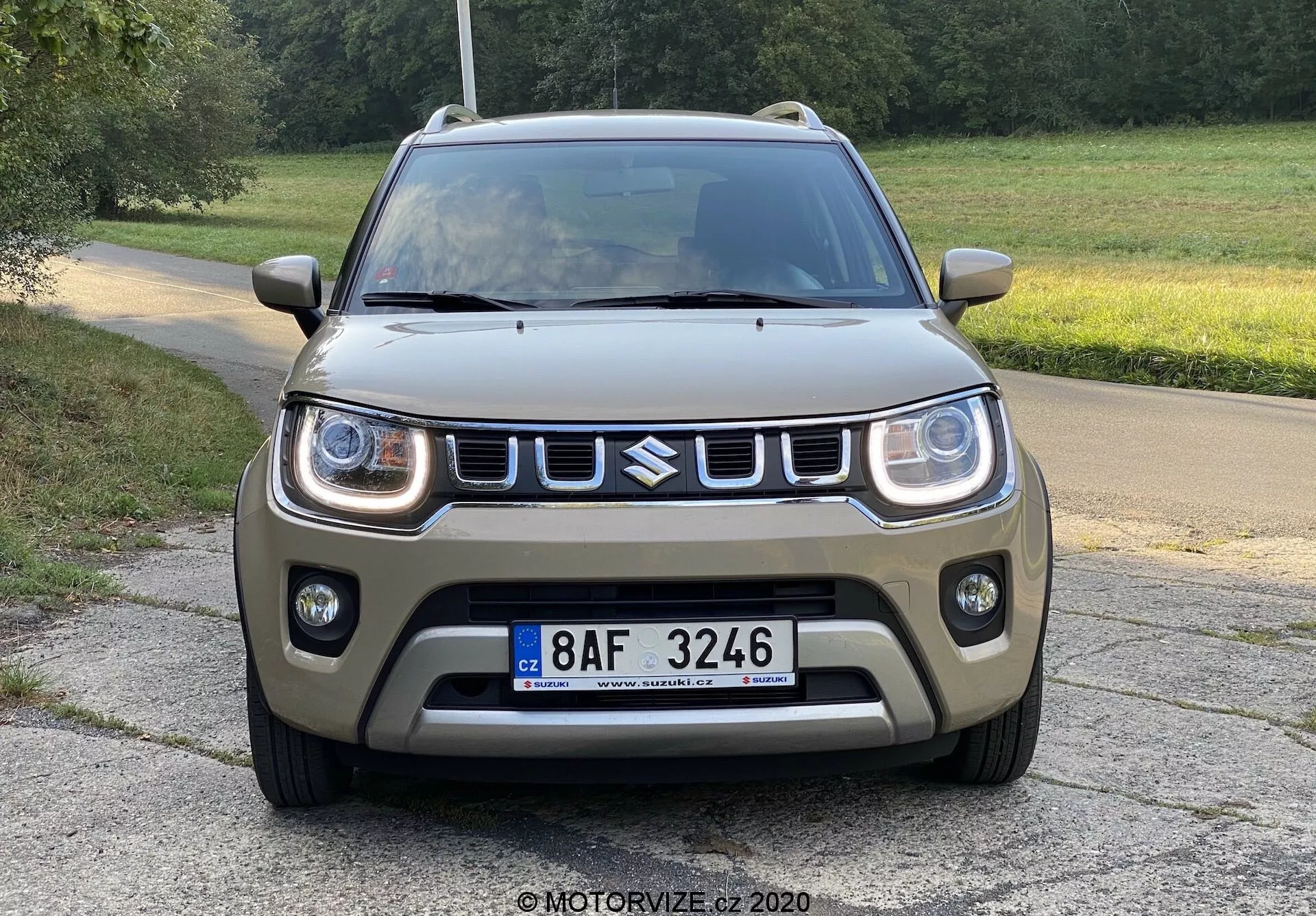 Přední pohled na kompaktní SUV Suzuki Ignis 2020 na dlážděné cestě s trávníkem v pozadí, venkovské nebo parkové prostředí, pohled z nízkého úhlu zdůrazňující design přední části, obsahující LED denní svícení, výraznou mřížku s logem, robustní přední nárazník s mlhovkami, matný zelenohnědý lak