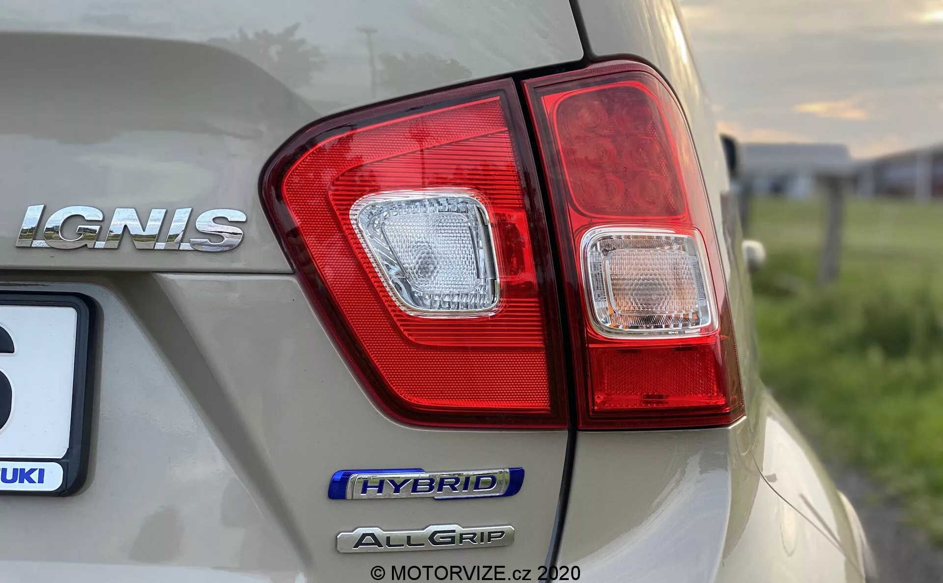 Detailní záběr zadního rohu Suzuki Ignis 2020, zaměřený na světelný klastr a označení modelu z nízké, šikmé perspektivy. Na pravé straně vozu jsou jasně viditelné označení 'IGNIS', 'HYBRID' a 'ALLGRIP', což zvýrazňuje hybridní technologii a systém pohonu všech kol. Vzhledem ke světle šampaňské barvě má auto lesklý povrch s odrazy prostředí a snímek zachycuje ostré detaily světel červené a čiré části, přičemž zachovává malou hloubku ostrosti.