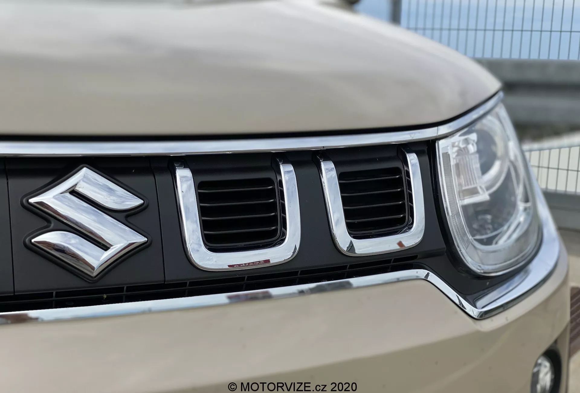 Zblízka přední mřížka a emblém vozu Suzuki Ignis 2020, zdůrazňující výrazné logo Suzuki, vertikálně orientované chromované sloty, levý světlomet s moderní LED nebo HID technologií a světlý odstín karoserie. Malá hloubka ostrosti zdůrazňuje detaily mřížky a loga, zatímco vzdálené hrany a pozadí jsou rozmazané.