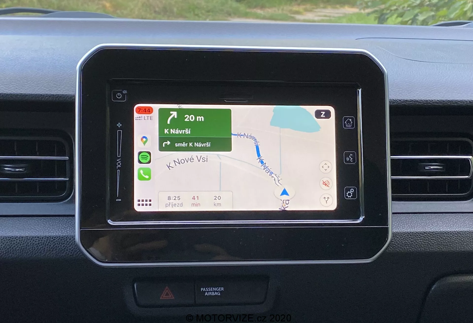 Detailní pohled na středový panel palubní desky Suzuki Ignis 2020, zaměřený na obdélníkovou obrazovku infotainment systému se zaoblenými rohy, zobrazující barevnou GPS navigační mapu se zelenou krajinou a modrými vodními plochami, udávajícími pokyny k odbočení, ETA, vzdálenost a rychlostní limity. Palubní deska má jednoduché designové prvky s horizontálními větracími otvory poblíž, kontrastujícími texturami materiálů a naznačuje dotykové nebo dálkové ovládání bez viditelného vnějšího kontextu.