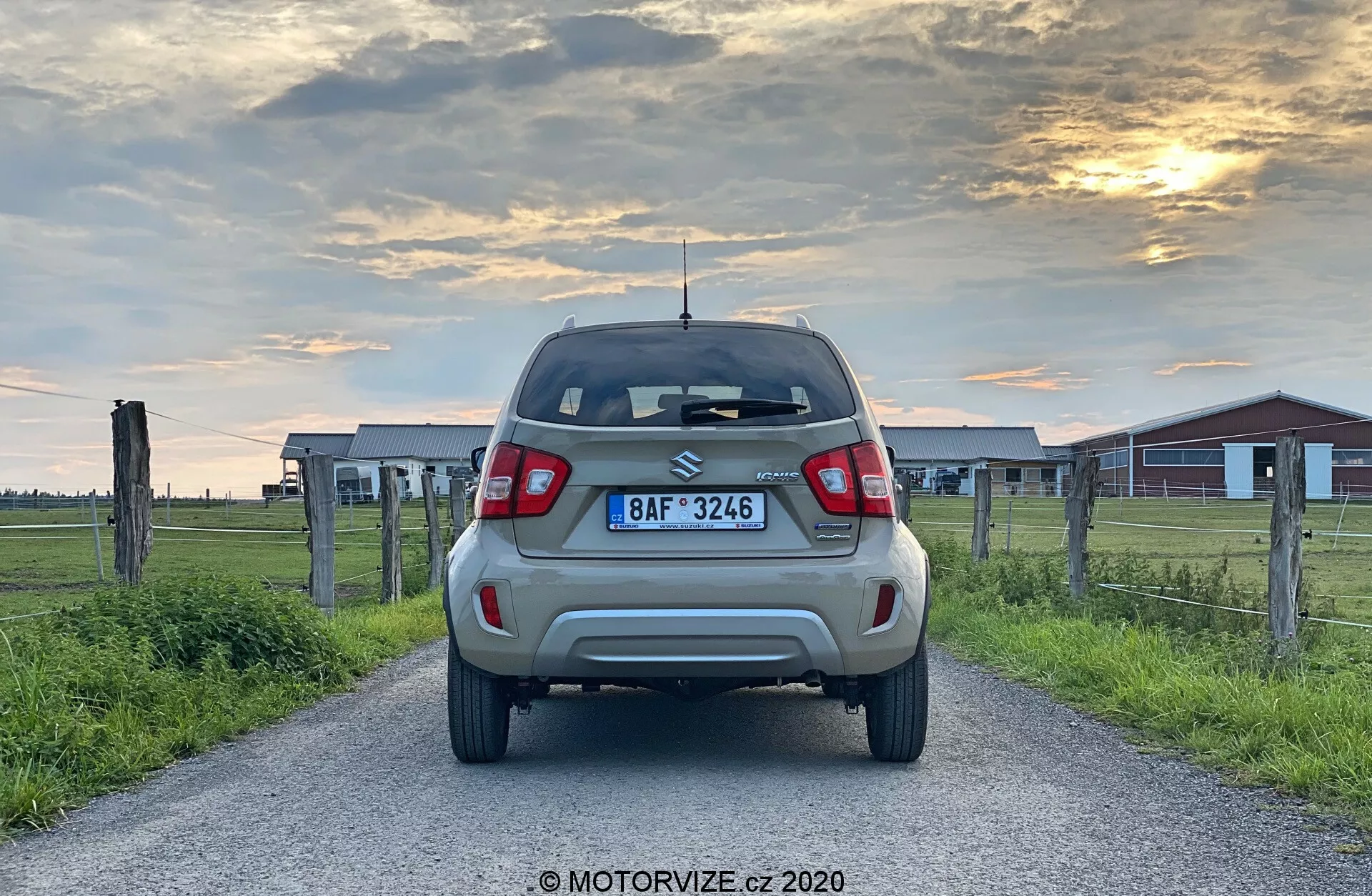 Zadní pohled na nehybný kompaktní SUV Suzuki Ignis 2020 1.2 Dualjet Hybrid Club na venkovské silnici za časného večera, ukazující moderní LED zadní světla, zadní spoiler, vysoko umístěné brzdové světlo, stěrač, zadní nárazník s parkovacími senzory a litá kola, na pozadí teplého západu slunce s dřevěnými ploty a hospodářskými budovami.