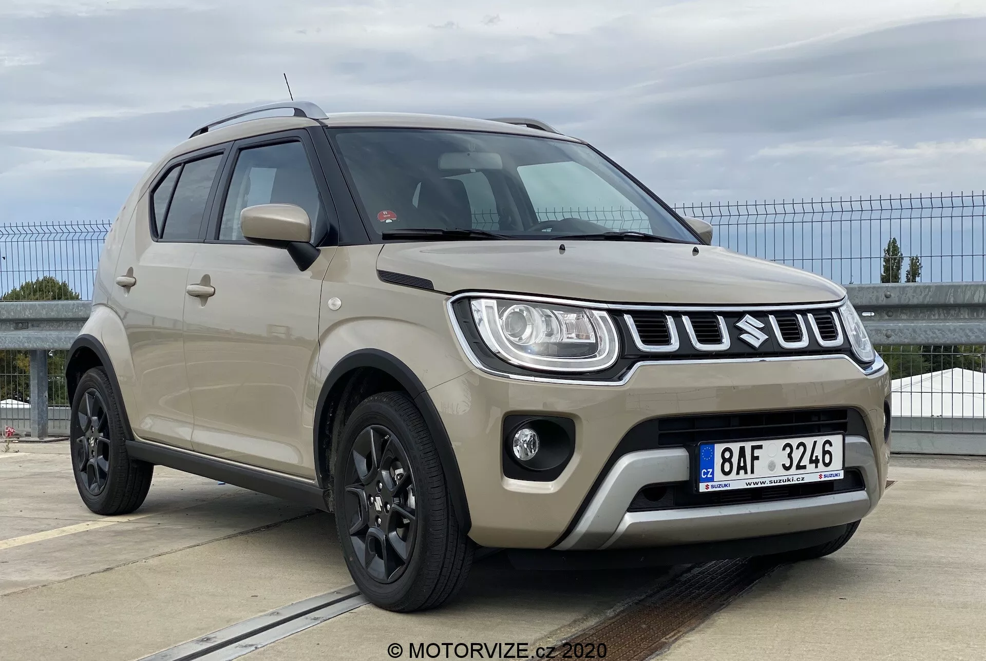 Obrázek zobrazuje Suzuki Ignis 1.2 Dualjet Hybrid Club z roku 2020 z nízkého třičtvrtinového předního úhlu, zdůrazňujícího jeho přední mřížku s vodorovnými chromovými lamelami, moderní LED nebo HID světlomety, mlhovky a sportovní tmavá litá kola. Robustní tělo vozu je natřeno tlumenou béžovou nebo světle khaki barvou s plastovými kryty na dolním okraji. Boční zrcátka odpovídají barvě karoserie a mají integrované směrovky. Střešní ližiny naznačují univerzálnost a viditelná je také přední registrační značka. Všímáme si tónovaných bočních oken a standardního čelního skla.