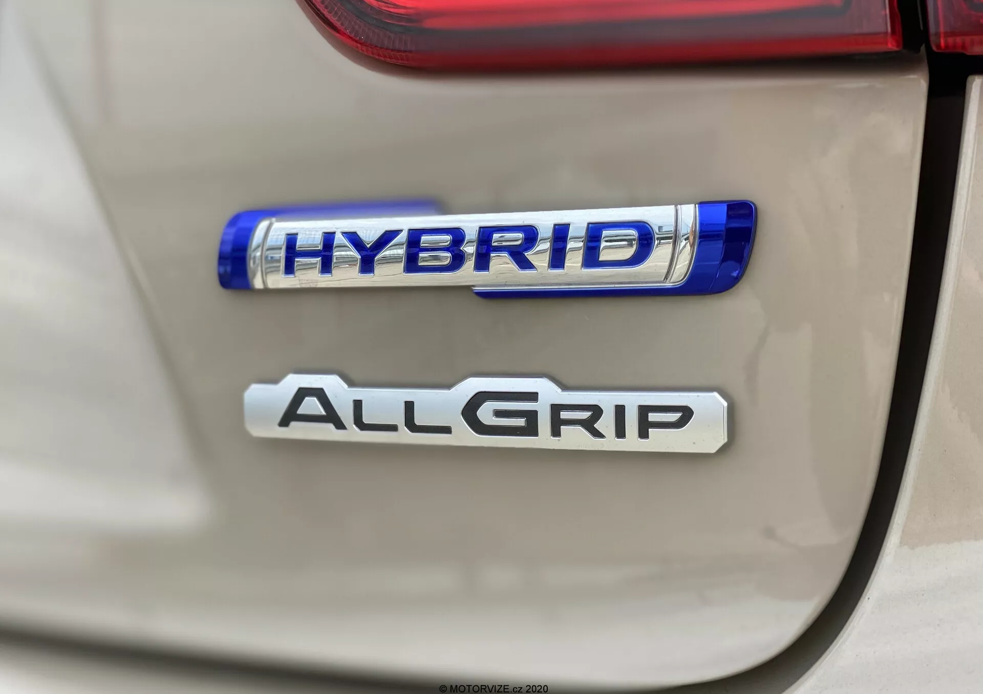 Detailní snímek zadních odznaků Suzuki Ignis 2020, zdůrazňující funkce 'HYBRID' a 'ALLGRIP'; jasný, přímý záběr s malou hloubkou ostrosti, zdůrazňující ekologicky šetrnou hybridní technologii a pokročilý systém pohonu všech kol; přirozené osvětlení s ostrými detaily a lehkými odlesky.