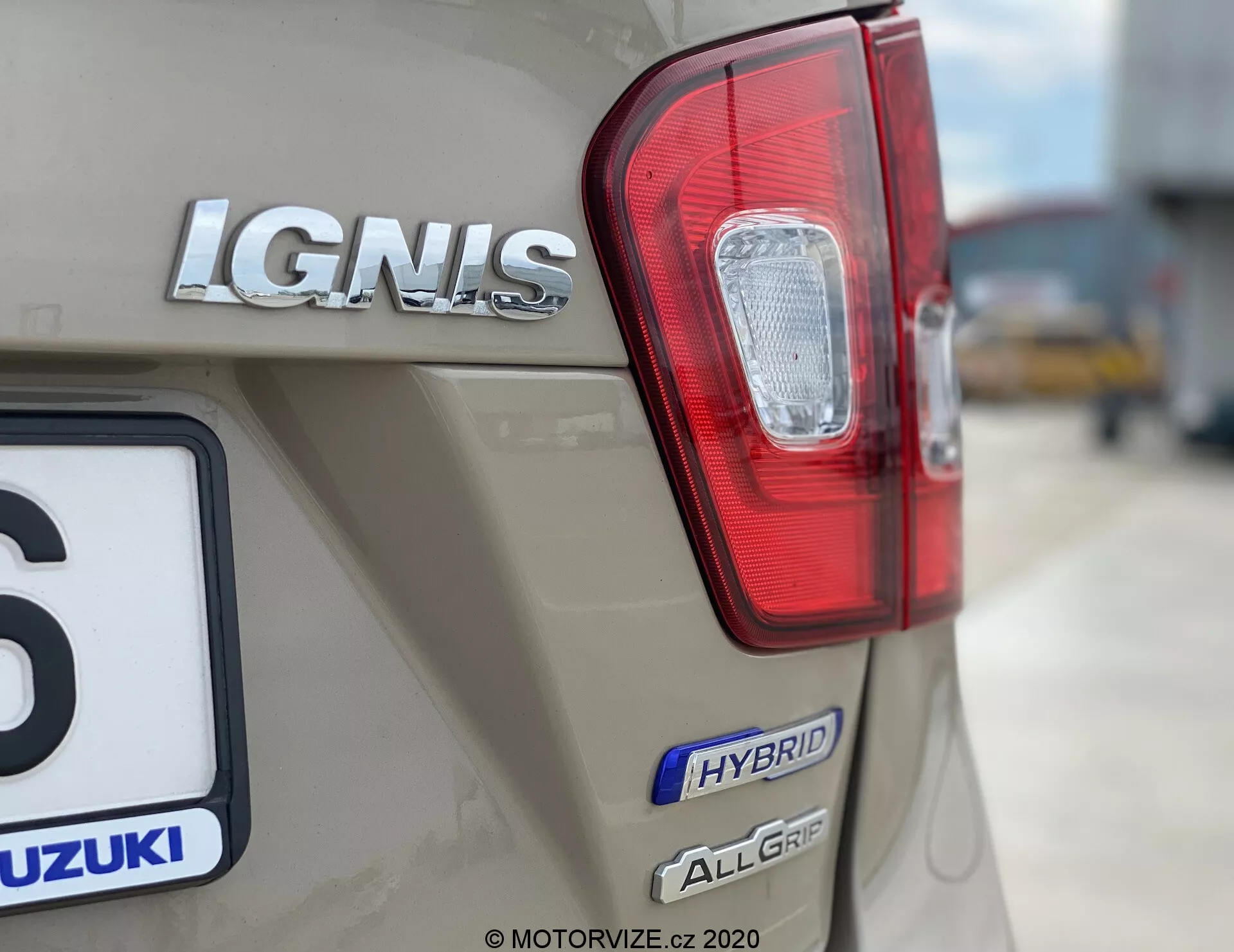 Detailní záběr na zadní část vozu Suzuki Ignis 1.2 Dualjet Hybrid Club z roku 2020, který zvýrazňuje pravé zadní světlo s červenými a čirými sekcemi, označení modelu 'IGNIS' v chromu, odznak 'HYBRID' a odznak 'ALLGRIP', což značí hybridní technologii a systém pohonu všech kol. Logo Suzuki je částečně viditelné nad registrační značkou, přičemž auto je béžové nebo světle hnědé metalízy. Pozadí je rozmazané, aby vynikl branding a technologické prvky vozidla.