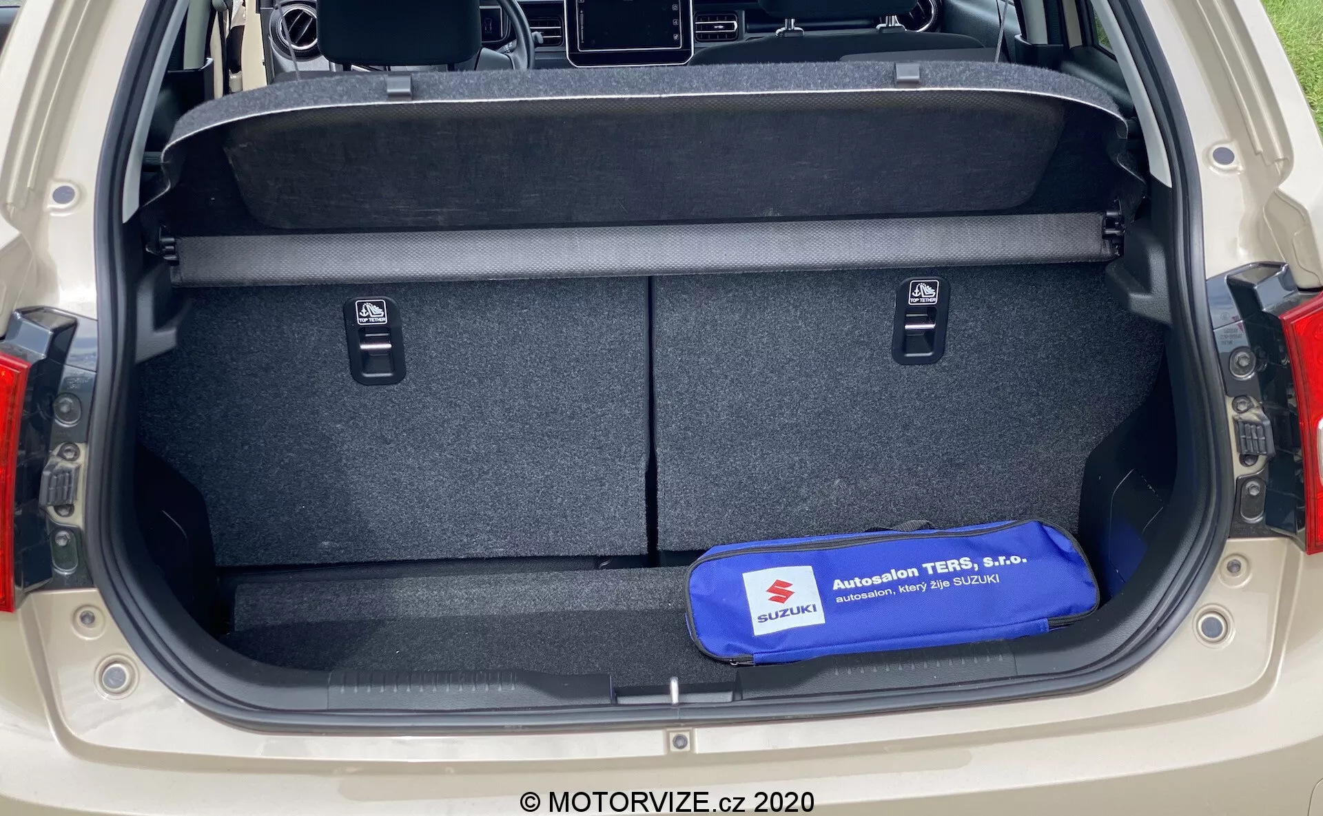 Kufr auta Suzuki Ignis 2020 z pohledu zezadu, zobrazující velký obdélníkový úložný prostor s kobercem na podlaze a stěnách, malé zapuštěné prostory pro předměty, táhla pro sklápění zadních sedadel, modrá bezpečnostní nebo nástrojová sada na podlaze, viditelný moderní design zadních světel po stranách kufru.