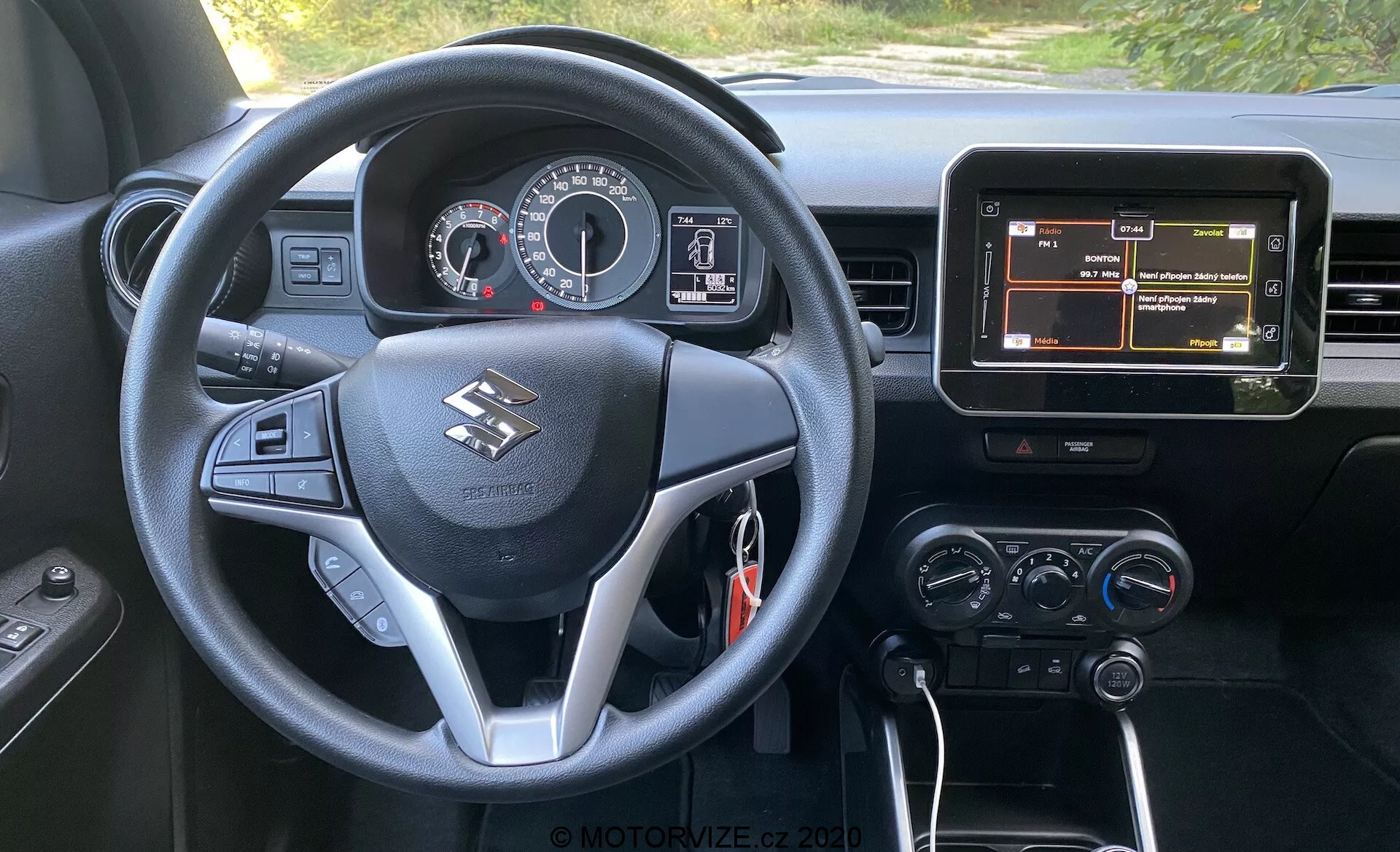 Pohled na interiér z místa řidiče Suzuki Ignis 1.2 Dualjet Hybrid Club 2020, zobrazující multifunkční volant s ovládacími prvky pro audio, telefon a informace o vozidle, přístrojovou desku s analogovými ukazateli a centrálním digitálním displejem, centrální infotainmentový displej zobrazující navigaci a ovládání klimatizace. Vozidlo má automatickou převodovku, což je naznačeno písmenem 'P' na řadicí páce, což naznačuje, že je zaparkováno, a černý interiér se stříbrnými akcenty.