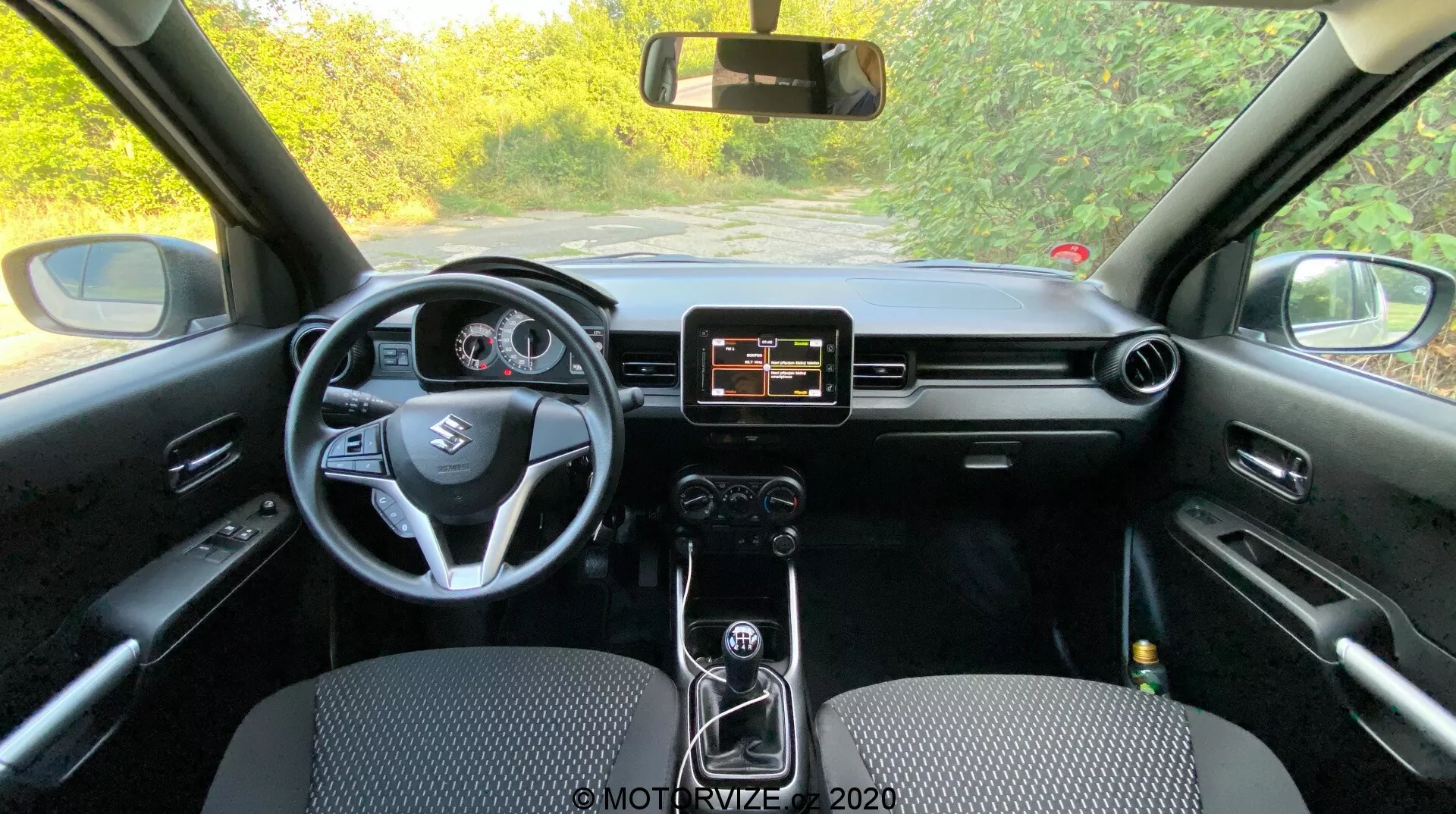 Pohled na interiér Suzuki Ignis 2020 zezadu ukazující palubní desku, volant, infotainment systém a přístrojový panel. Středová konzola s manuálním řazením, černým interiérem s různými texturami a materiály.