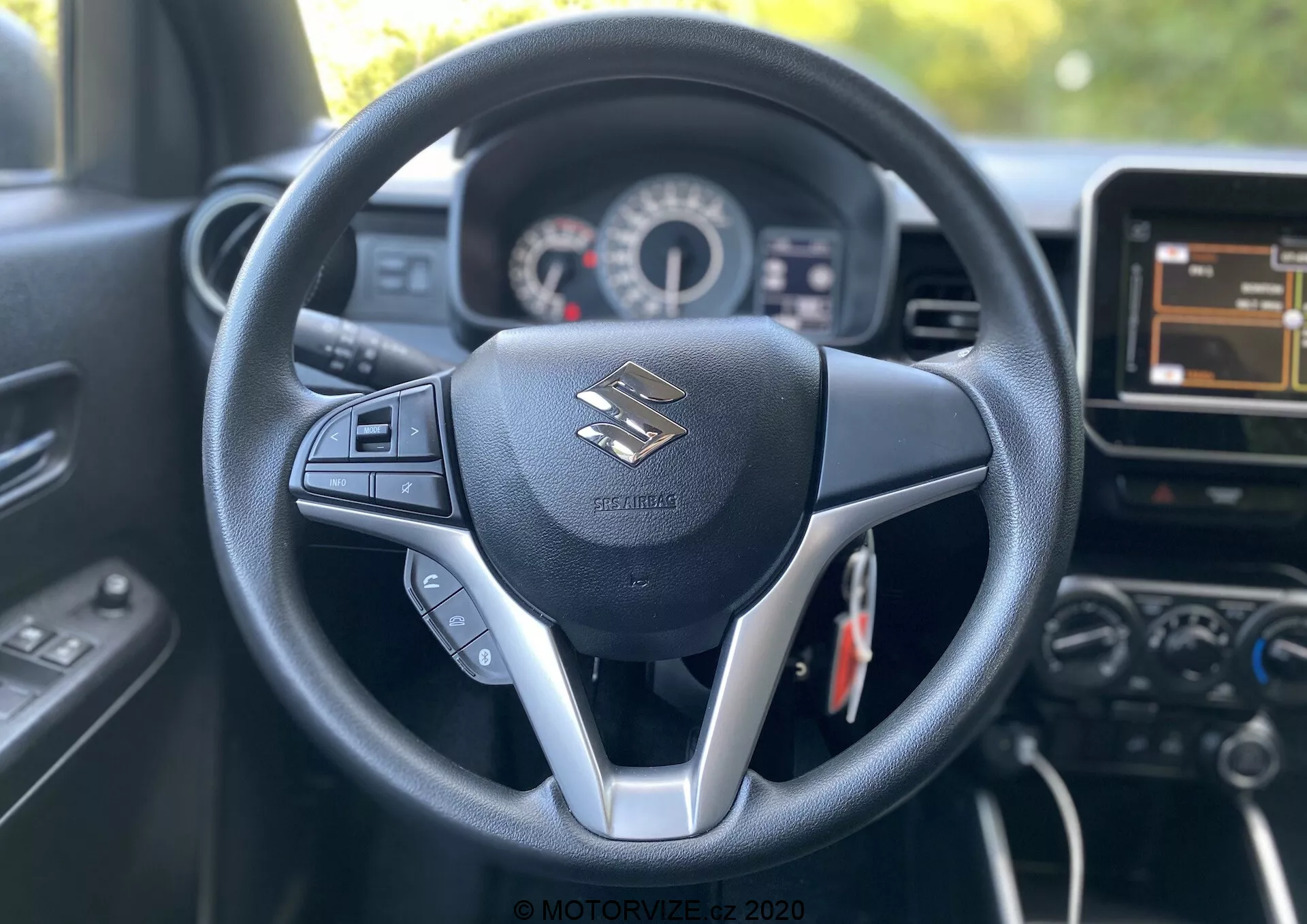 Interiér vozu Suzuki Ignis 2020 z pohledu řidiče, zobrazující centrální volant s trojpaprskovým designem a stříbrným akcentem. Volant má ovládací prvky pro audio a pravděpodobně i tempomat. Před ním se nachází přístrojová deska zobrazující tachometr a otáčkoměr s malým digitálním displejem. Snímek také zachycuje část středové konzole, včetně dotykového infotainment systému a dalších ovládacích prvků. Interiér působí moderně s černým barevným schématem, syntetickou kůží a plastovými prvky, které kladou důraz na pohodlí řidiče a technologie.