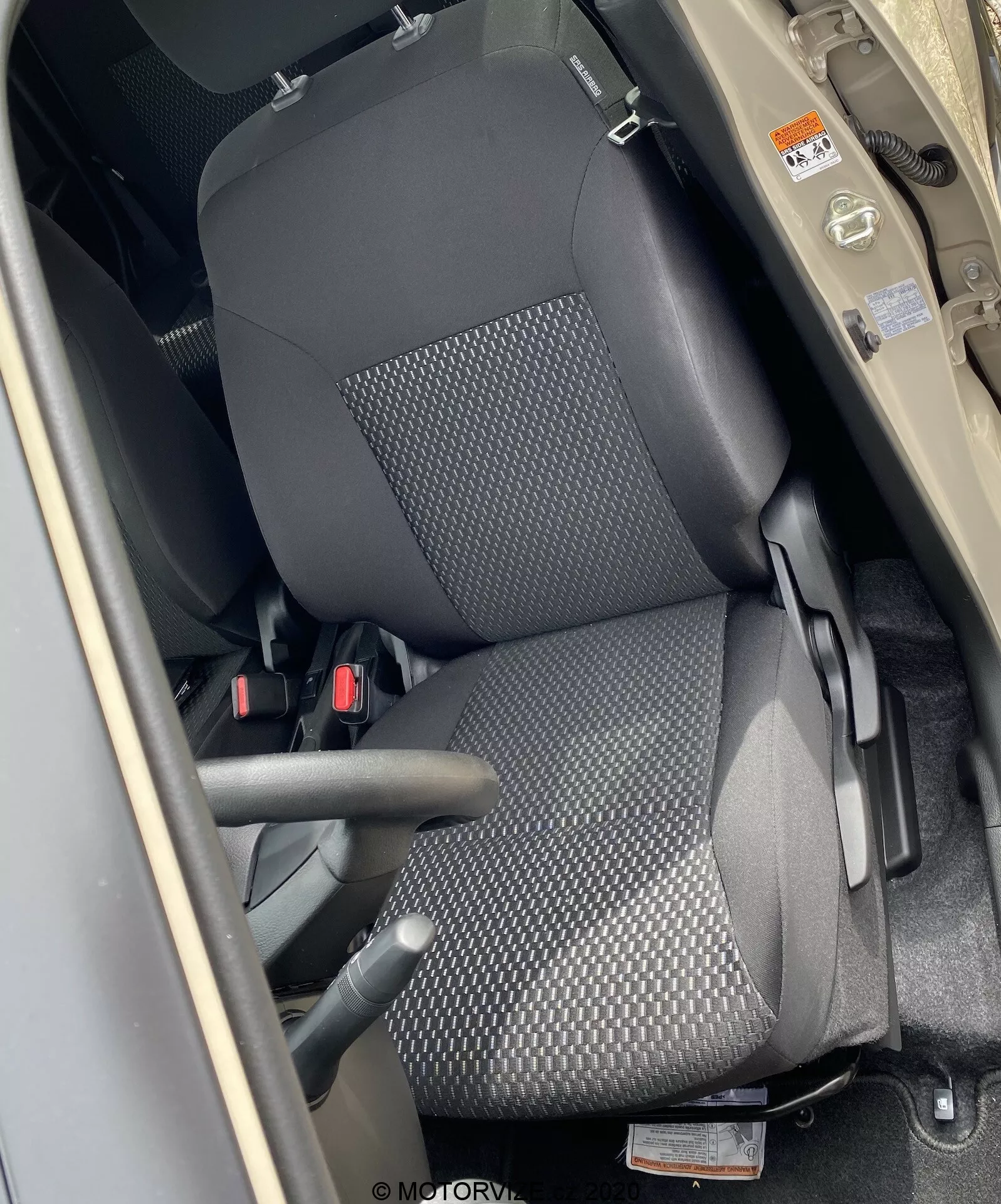 Detailní pohled shora na přední sedadlo spolujezdce v Suzuki Ignis 2020, ukazující opěrku hlavy, opěradlo a sedací část. Záběr je z perspektivy osoby stojící vedle otevřených dveří umístěných kolem B-sloupku. Sedadlo je čalouněno černou texturovanou látkou s tečkovaným vzorem a má ruční nastavení. Je viditelný tříbodový bezpečnostní pás, béžový či světle hnědý interiér tvoří kontrast s černým sedadlem.