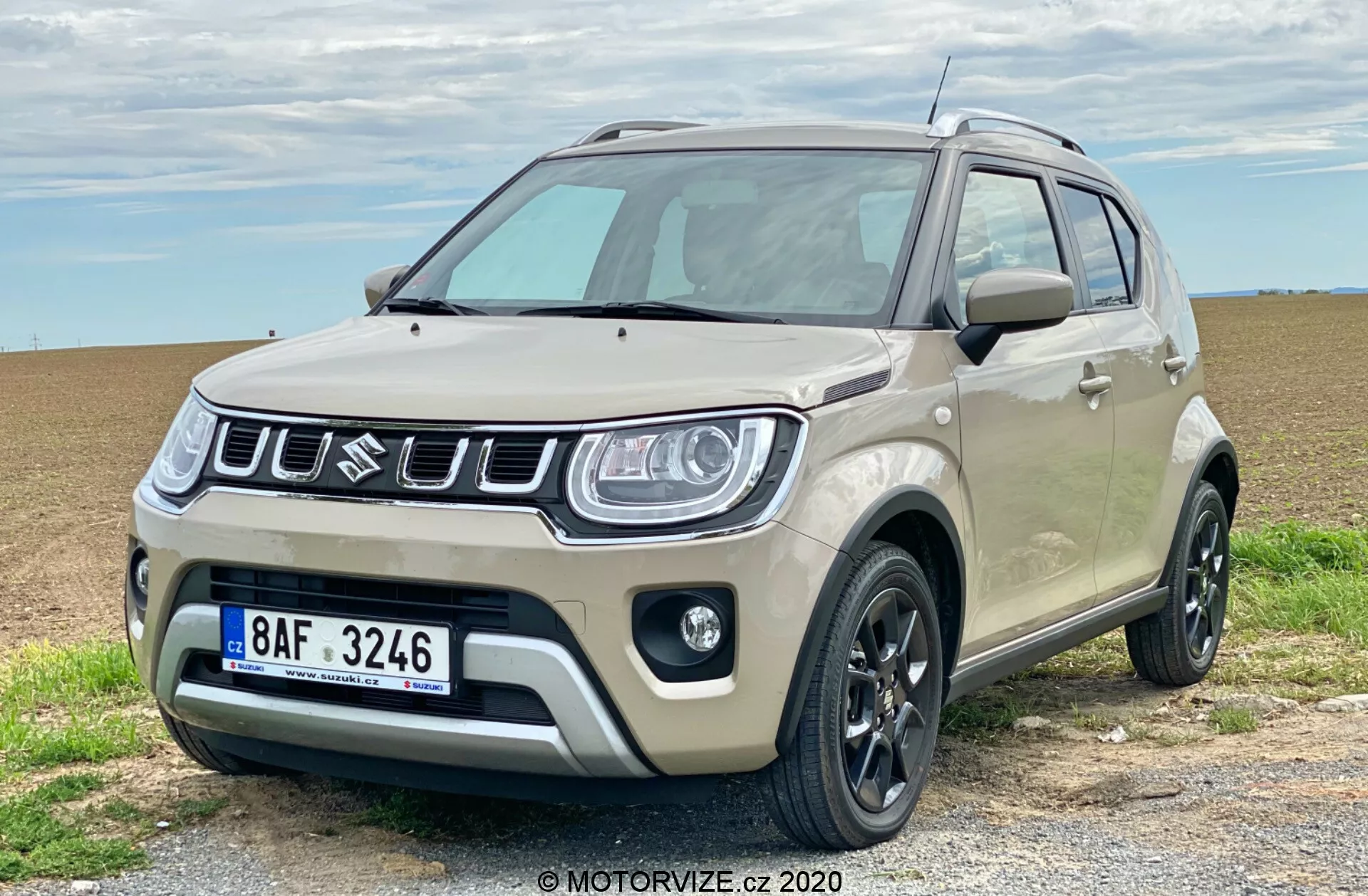 Kompaktní SUV Suzuki Ignis 1.2 Dualjet Hybrid Club 2020 je zaparkované na štěrkové cestě s otevřeným polem v pozadí, zobrazující třičtvrteční přední pohled. Má dvoubarevný nástřik s hlavní barvou světle béžovou a černými akcenty na bočních zrcátkách, střešních ližinách a náraznících. Výrazná přední mřížka má vertikální lamely s centrálním znakem značky. Moderní světlomety s LED denními světly, černá litá kola, plastové obložení, mlhovky a vysoká světlá výška jsou výraznými prvky. Vozidlo vyzařuje jak městský šarm, tak lehké off-road schopnosti.