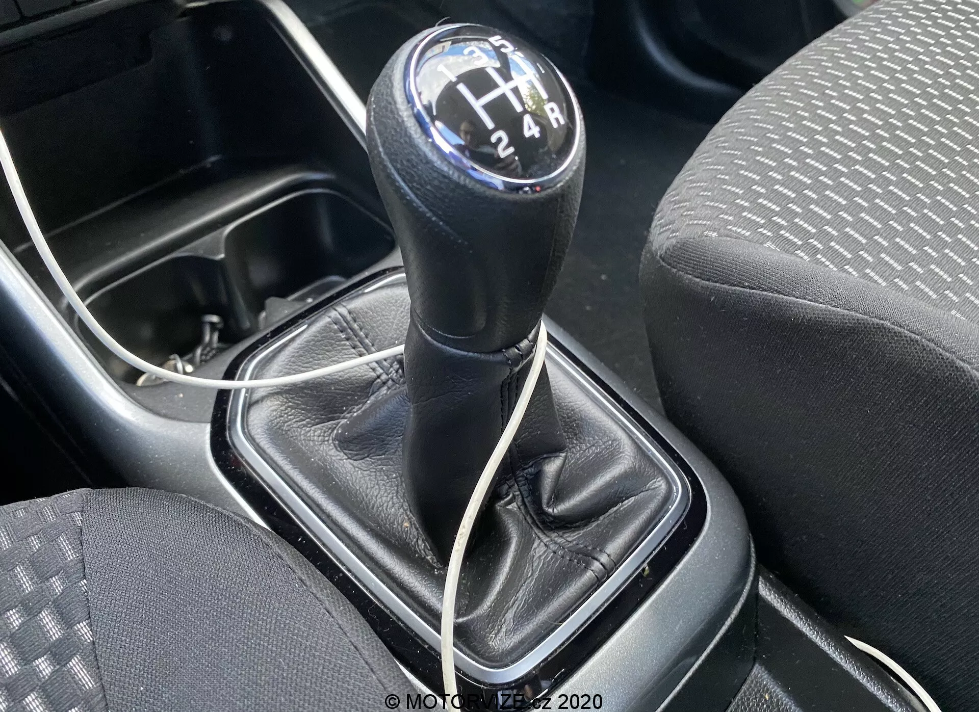 Detailní pohled na řadicí páku manuální převodovky vozu Suzuki Ignis 1.2 Dualjet Hybrid Club 2020, snímek je pořízen z horního úhlu a zaměřuje se na kulatý černý řadicí knoflík s bílými číslicemi v rozvržení 6stupňové převodovky do tvaru H a couvacího stupně. Řadicí páka je v neutrální poloze. Kolem páky je černá kožená manžeta s viditelným šitím a plastová konzole stříbrné nebo šedé barvy. Vlevo je vidět část sedadla s látkou s motivem teček a linek, a pod řadicí pákou částečně viditelný bílý kabel naznačující moderní konektivitu, například nabíjecí port. Vlevém dolním rohu je také vidět okraj držáku na nápoje nebo malé úložné přihrádky, což zdůrazňuje praktický design středové konzole vozu.