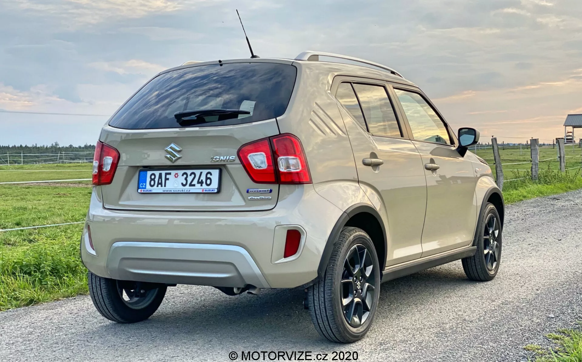 Zadní třičtvrtinový pohled na 2020 Suzuki Ignis 1.2 DualJet Hybrid Club, moderní kompaktní SUV, umístěné na venkovské cestě nebo travnatém terénu s rozlehlým nebem při svítání nebo soumraku. Viditelné prvky zahrnují LED zadní světla, výrazný zadní nárazník se senzory nebo reflektory, zadní stěrač, vysoko umístěnou zastavovací lampu, černá litá kola s pneumatikami s nízkým profilem, zrcátka se směrovými světly, stoupající linii oken, anténu na střeše, střešní ližiny a emblémy značky. Kondice vozidla a tlumené barevné schéma jsou zvýrazněny měkkým osvětlením.