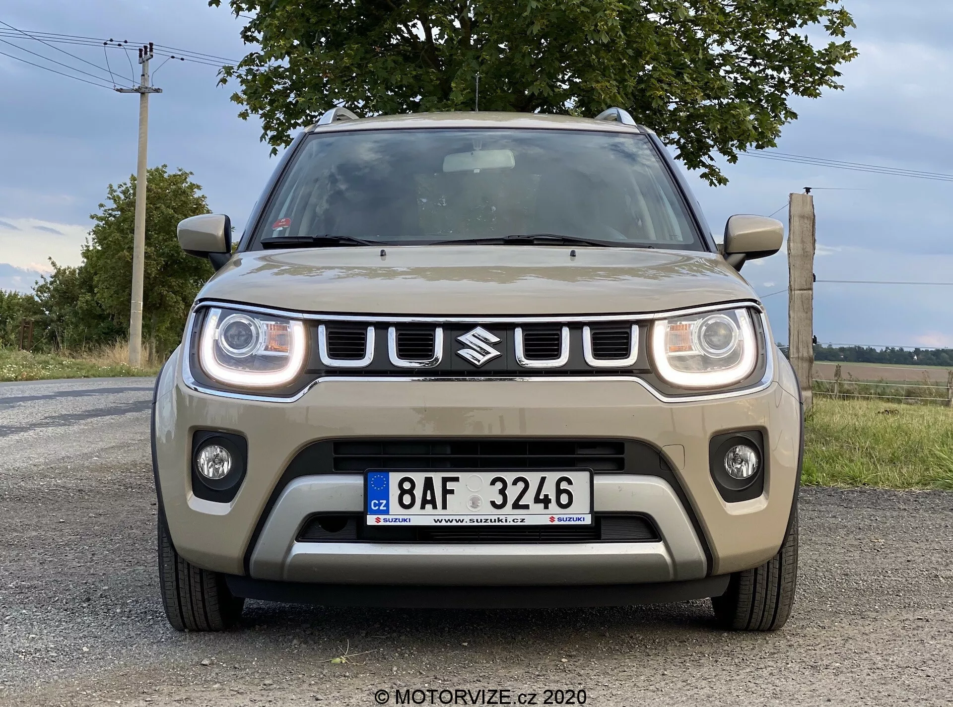 Čelní pohled na Suzuki Ignis 2020 (Suzuki Ignis 1.2 Dualjet Hybrid Club) pořízený z mírného nízkého úhlu, zobrazujícího přední části včetně mřížky s emblémem, obdélníkových LED světlometů, mlhovek a centrálního nasávacího otvoru na dlážděné silnici s přírodní krajinou a zataženou oblohou v pozadí.