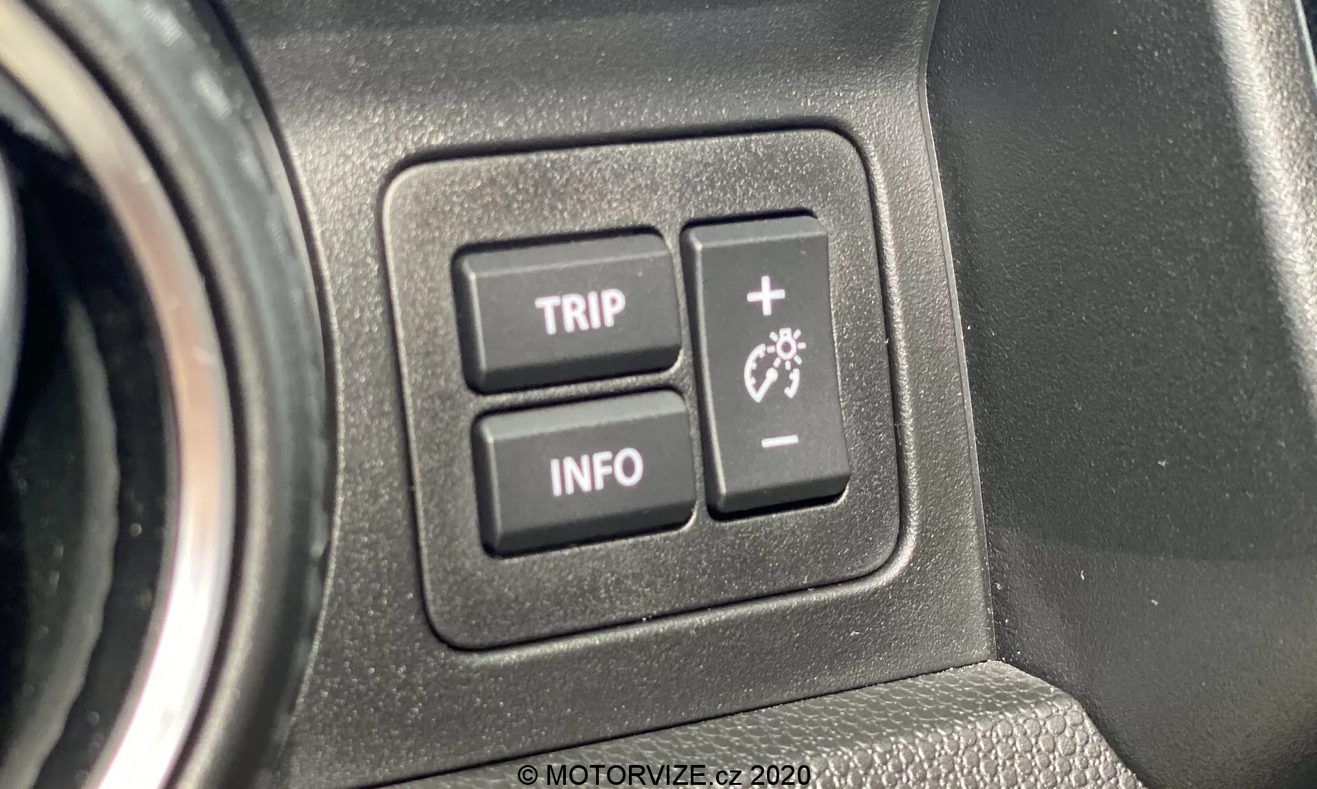Detailní pohled na ovládací prvky interiéru modelu Suzuki Ignis 2020. Obrázek se zaměřuje na dvě tlačítka označená 'TRIP' a 'INFO,' která jsou součástí palubního počítače vozidla, umístěná na texturovaném plastovém panelu. Tlačítko 'TRIP' pravděpodobně přistupuje k funkcím počítače cesty, zatímco tlačítko 'INFO' zobrazuje různé informační prvky o vozidle. Detailní záběr zdůrazňuje ergonomický design a uživatelsky přívětivé rozhraní, což zvýrazňuje moderní technologickou integraci ve vozidle.