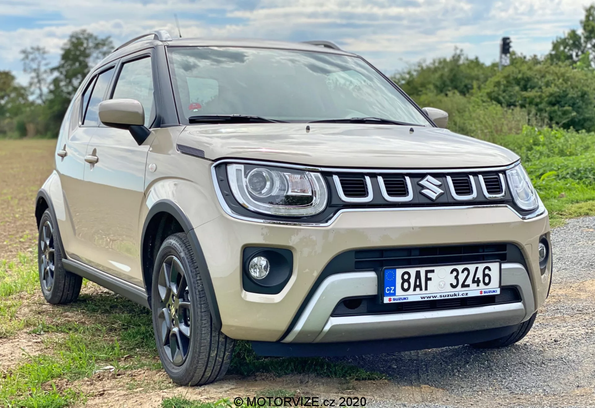 Moderní subkompaktní crossover SUV béžové barvy, pravděpodobně model Suzuki Ignis 2020, zaparkovaný na štěrkovém povrchu v polovenkovské oblasti. Vozidlo, zachycené z nízkého úhlu ve frontální tříčtvrteční perspektivě, demonstruje svůj výrazný design přední části včetně nápadné mřížky, moderních světlometů a mlhových světel. Vícepaprsková litá kola, vyšší světlá výška, ochranné lišty a integrované směrovky na zrcátkách podtrhují jeho současné a odolné prvky. Mělká hloubka ostrosti s přirozeným osvětlením soustředí pozornost na vozidlo.