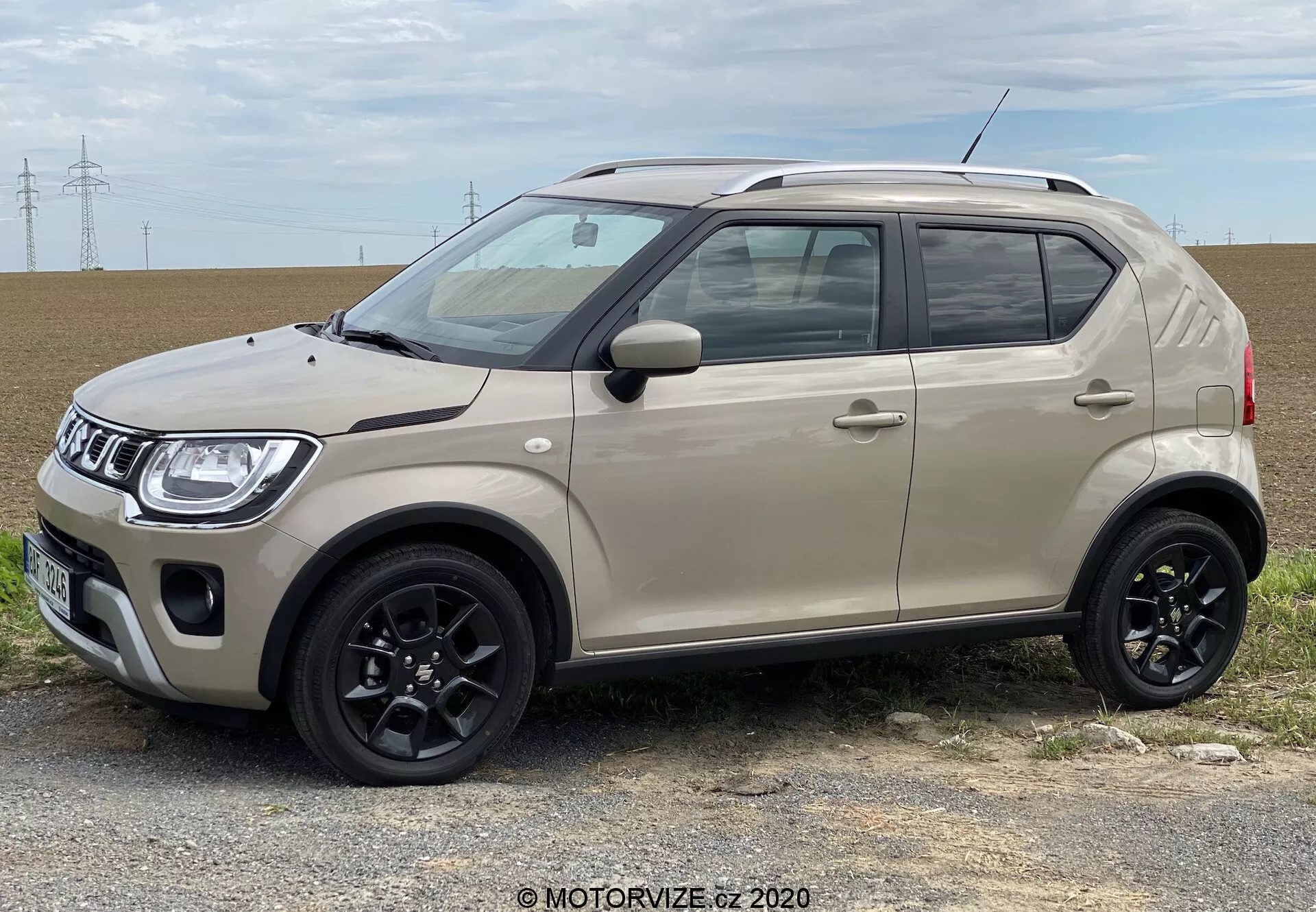 Obrázek kompaktního SUV z roku 2020 zaparkovaného na rovné ploše s přírodní krajinou a elektrickými vedeními v pozadí. Auto je viděno z bočního profilu s mírným náklonem dopředu, ukazuje moderní přední mřížku s geometrickým vzorem, ostré světlomety, dvoubarevnou karoserii světle béžové a černou střechu, černé plastové obložení kolem oblouků kol a bočních prahů, černá zpětná zrcátka s možnými směrovkami a černá vícepaprsková litá kola vhodná pro městskou jízdu a mírný terén. Krátká anténa je také viditelná směrem dozadu.