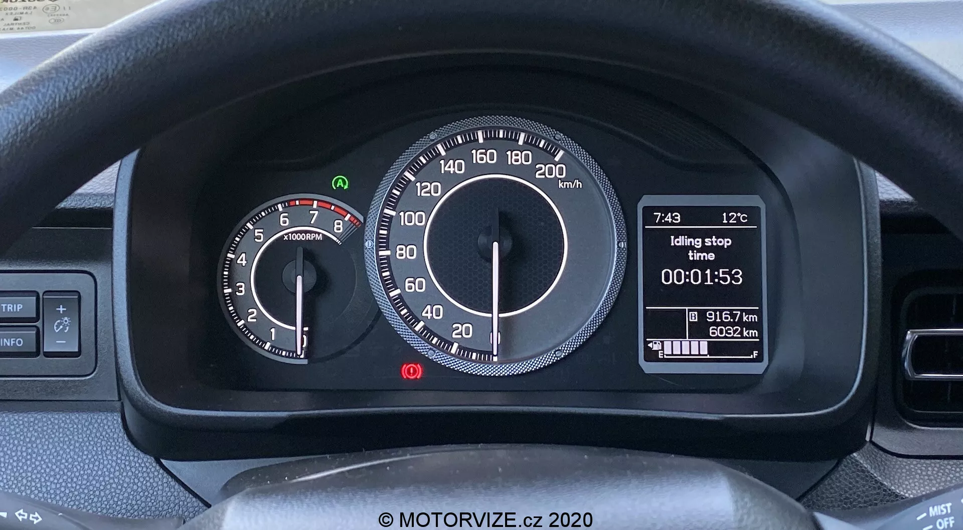 Záběr interiéru vozu Suzuki Ignis 2020 zaměřený na přístrojovou desku za volantem. Viditelná je horní část volantu a ovladače pro směrovky a stěrače. Přístrojová deska kombinuje analogové a digitální displeje: vlevo analogový otáčkoměr s červenou zónou při 6500 ot/min a vpravo analogový tachometr do 220 km/h. Centrální digitální displej ukazuje venkovní teplotu (12°C), čas (7:43), funkci 'Idling stop', časovač (00:01:53) a počítadlo kilometrů (91672 km). Moderní, uživatelsky přívětivý design pro trhy s metrickým systémem.