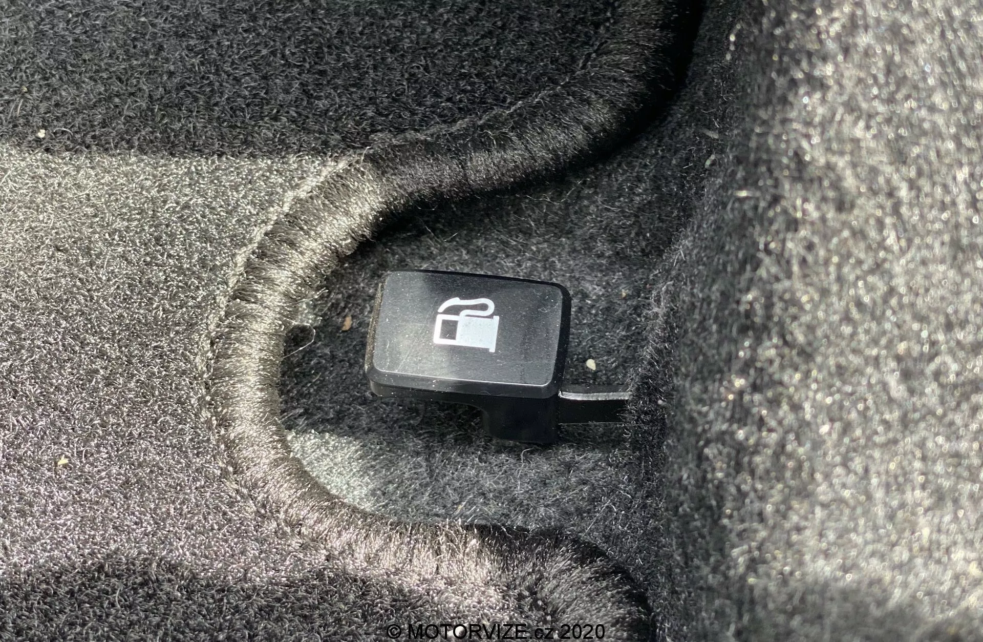 Detailní pohled na tlačítko uvolnění kufru u Suzuki Ignis 1.2 Dualjet Hybrid Club 2020, umístěné v černém, kobercovým interiéru. Tlačítko se symbolem auta s otevřeným kufrem nabízí pohodlný přístup k otevření kufru zevnitř vozidla. Záběr ze šikmého pohledu nad sedadlem zdůrazňuje dobře udržované textury a praktický design tohoto prvku interiéru vozidla.