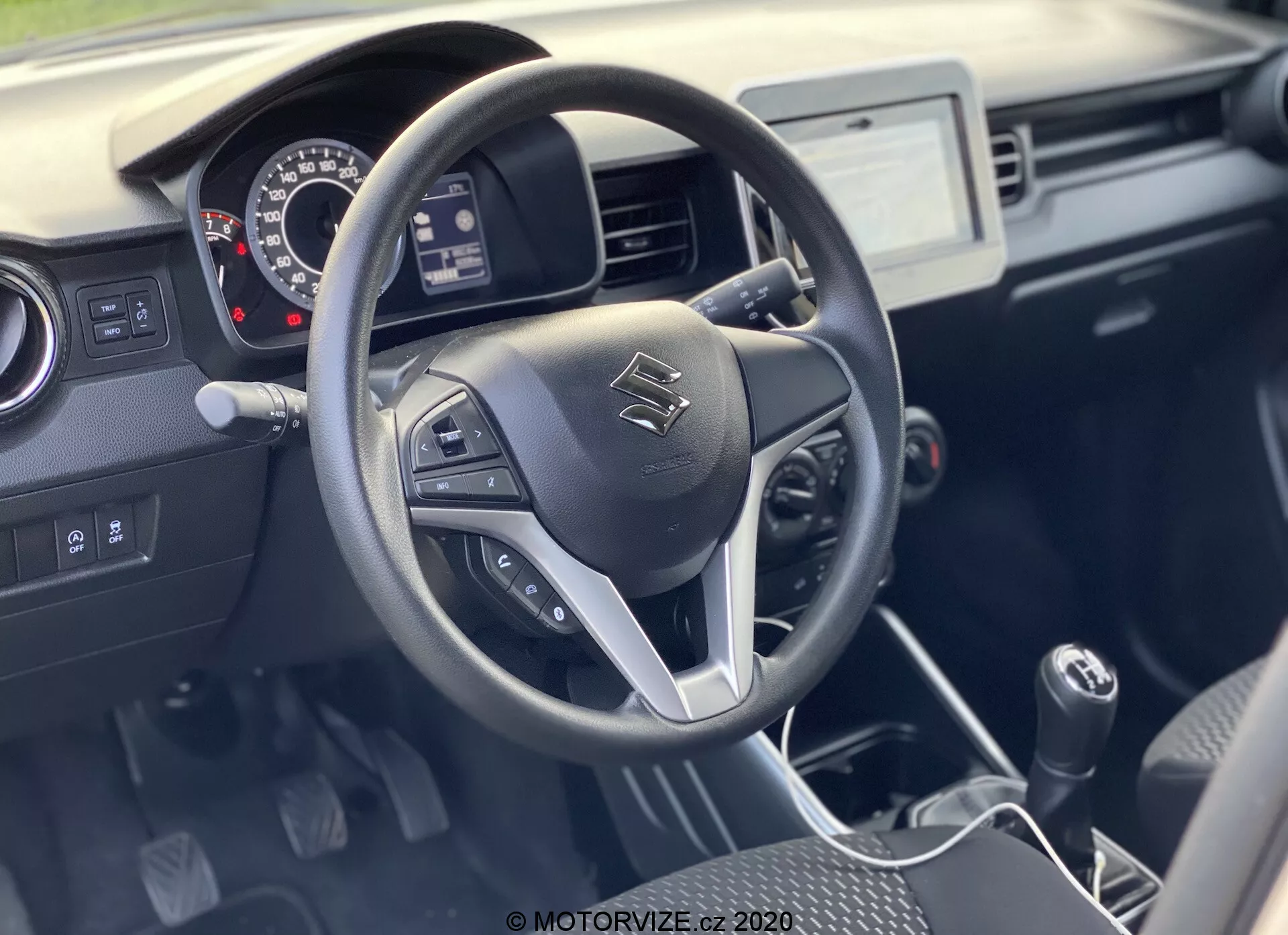 Interiér modelu Suzuki Ignis 1.2 Dualjet Hybrid Club z roku 2020 s tříramenným volantem a integrovanými ovládacími prvky pro audio, hands-free volání a tempomat. Palubní deska obsahuje analogové měřidla, středový infotainment displej s dotykovým ovládáním a řadicí páku manuální převodovky. Praktický dvoubarevný interiér klade důraz na funkčnost s odolnými materiály a dobrou viditelností za denního světla.