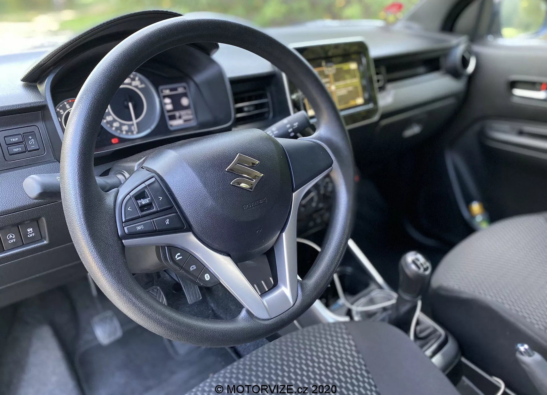 Pohled na interiér z pohledu řidiče vozu Suzuki Ignis 2020 (1.2 Dualjet Hybrid Club) se zaměřením na moderní tříramenný volant s multifunkčními tlačítky. V centru volantu je znak Suzuki, obklopený stříbrnými akcenty a kvalitním prošíváním. Za volantem je částečně viditelný přístrojový panel s digitálním displejem a analogovými číselníky zobrazujícími rychlost, otáčky a stav vozidla. Na středovém panelu je infotainmentový systém, který pravděpodobně nabízí audio, navigaci a nastavení vozidla. Na středové konzoli je řadicí páka manuální převodovky, což naznačuje manuální převodovku. Interiér je převážně černý s texturálními prvky, sedačky s ornamentálním vzorem, naznačující důraz na estetiku a pohodlí.