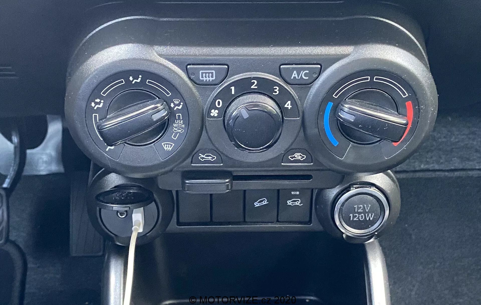 Detailní záběr na středovou konzoli Suzuki Ignis 2020, zaměřený na jednotku klimatizace se třemi hlavními knoflíky pro teplotu, rychlost ventilátoru a směr proudění vzduchu. Regulátor teploty má modré a červené indikátory pro chladné a teplé nastavení. Nad středovým knoflíkem ventilátoru je tlačítko 'A/C', recirkulace vzduchu v kabině a tlačítka pro přední odmlžování. Pod ovladači je umístěna zásuvka 12V 120W, což zdůrazňuje praktický a uživatelsky přívětivý design s manuálními přepínači.