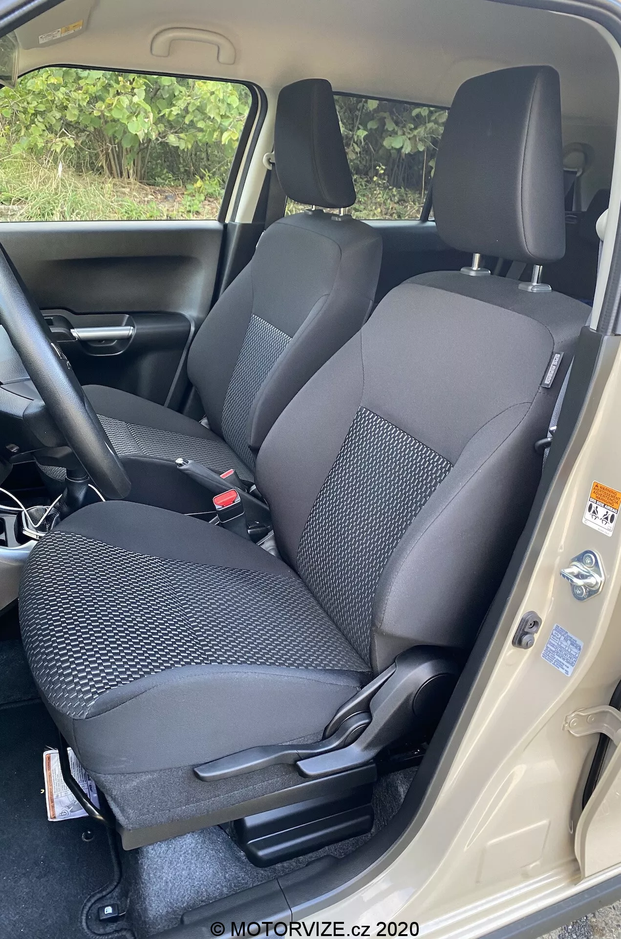 Profesionální fotografie interiéru Suzuki Ignis 2020 (Suzuki Ignis 1.2 Dualjet Hybrid Club), zaměřená na přední sedadlo spolujezdce. Pořízena z výšky, kamera zachycuje jasný pohled s otevřenými dveřmi vozu. Dvoubarevné látkové čalounění sedadla má vzorovaný design pro odolnost. Nastavení sedačky je manuální, s opěrkou hlavy, bezpečnostním pásem a viditelnými pákami na ladění. Viditelná je také část středové konzoly a vnitřní panel dveří s regulačními štítky. Tmavý koberec a světlý práh dveří zdůrazňují kontrast designu. Obrázek je připsán MOTORVIZE.cz 2020.