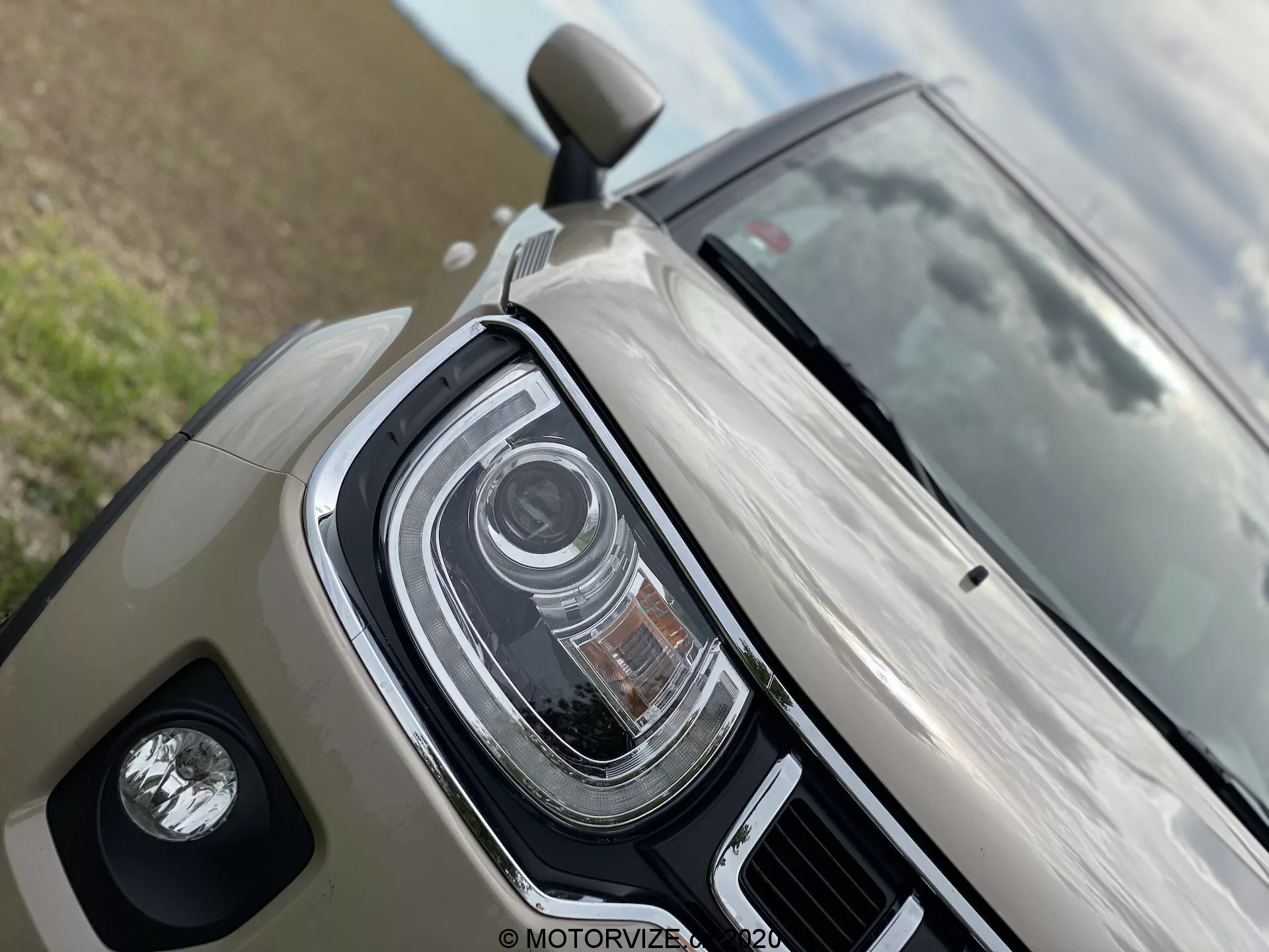 Detailní záběr na Suzuki Ignis 2020, zdůrazňující levý přední kvadrant. Obsahuje LED světlomety s více oddíly, které mohou být adaptivní světlomety nebo automatická dálková světla. Ukazuje lesklý béžový/světle šedý lak a kryt mlhovky. Částečný pohled na boční zrcátko naznačuje pokročilou techniku, jako je signalizace v bočních zrcátkách nebo sledování mrtvého úhlu.