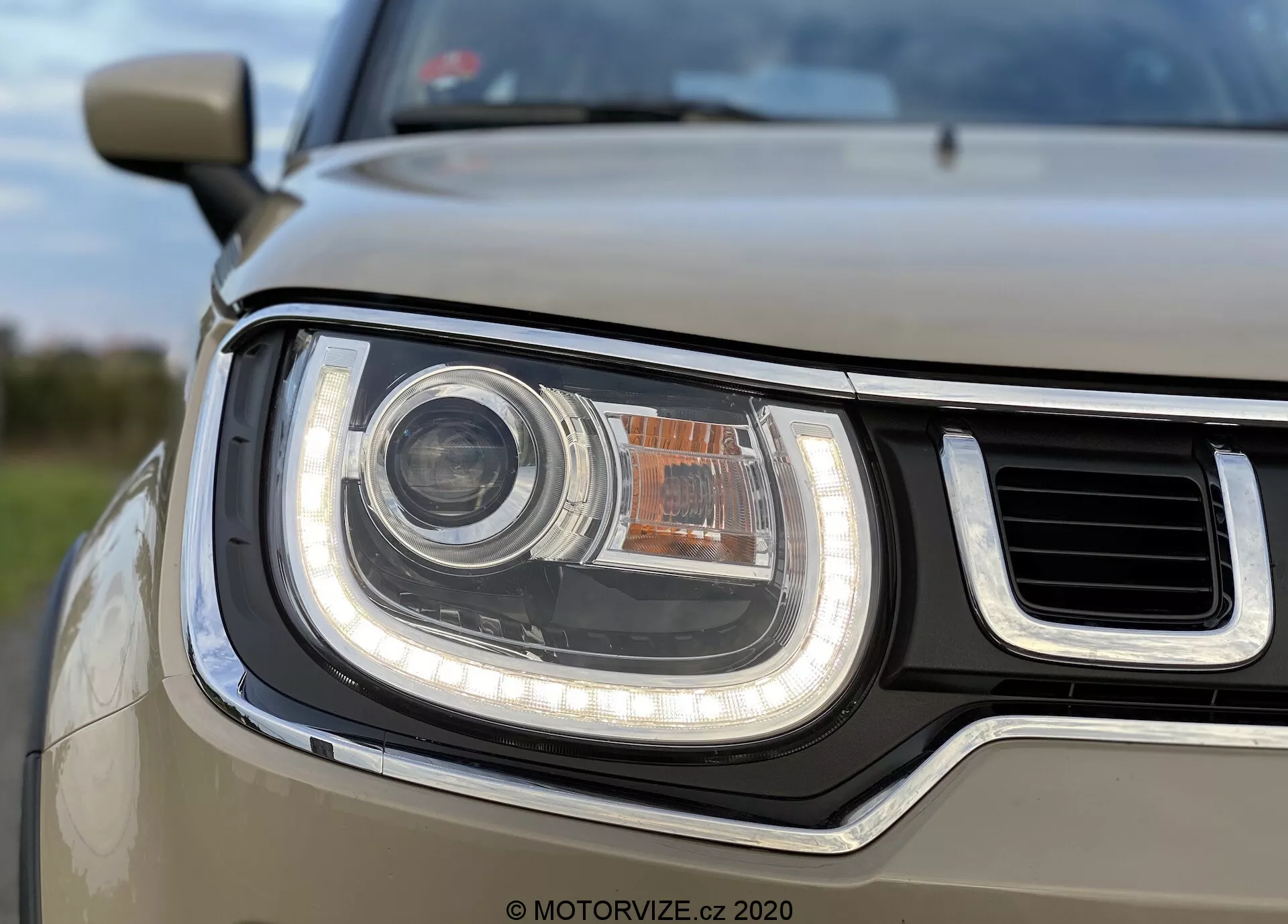 Pohled zblízka na světlovodné sestavení na přední levé straně Suzuki Ignis 2020, zobrazující kruhový hlavní světlomet pravděpodobně LED nebo HID, obklopený LED trubicovým denním svícením (DRL). Světlomet obsahuje menší jantarový směrový nebo parkovací světlo nahoře. Částečně viditelná mřížka má vodorovné tyče, se světlovodným designem tvořeným zakřivenými a rovnými hranami, což vytváří moderní a robustní vzhled. Hloubka ostrosti se zaměřuje na světlomet, zatímco pozadí a další části vozu jsou trochu rozmazané.