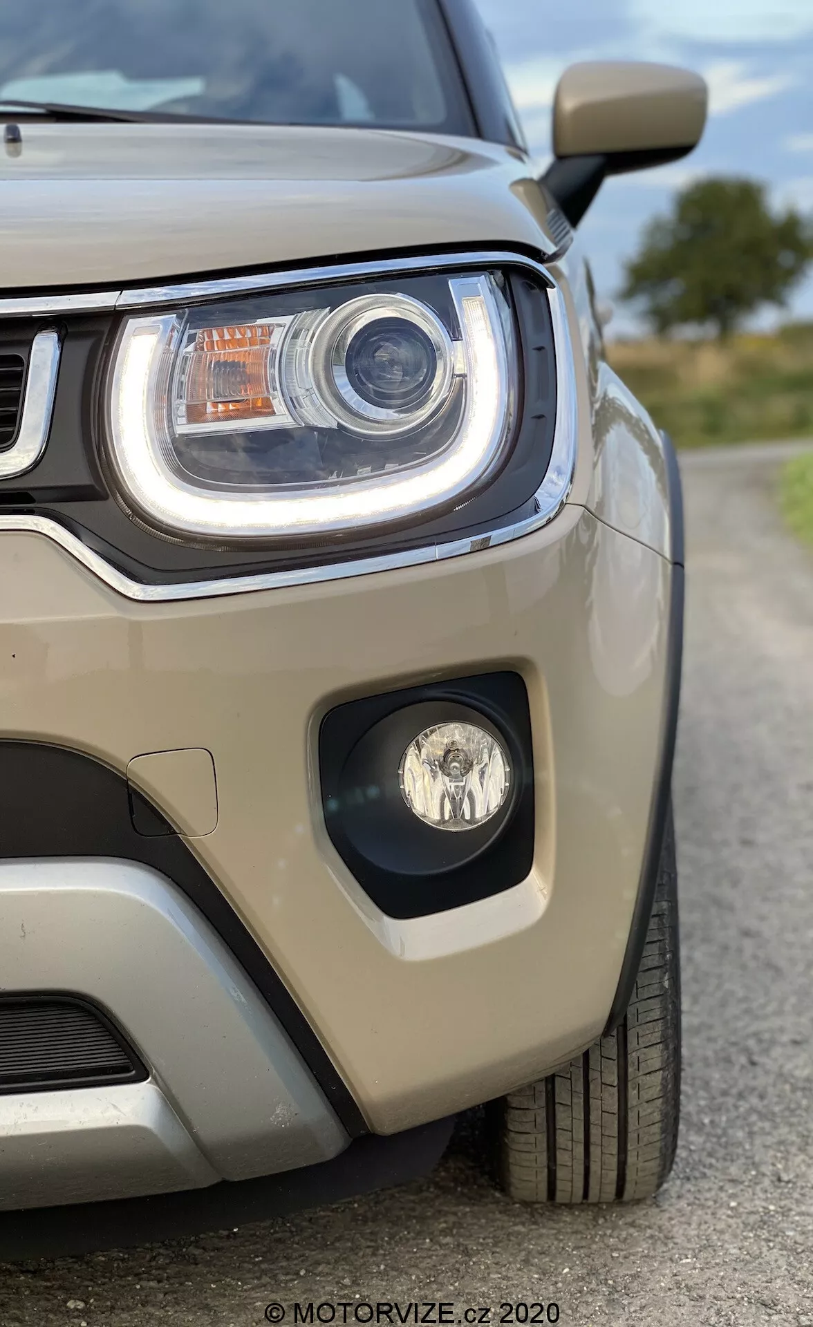 Detailní pohled zepředu vlevo na Suzuki Ignis 2020 s důrazem na světlomet a část mřížky. Obrázek pořízený z mírně nízkého úhlu zdůrazňuje agresivní linie světlometu. Obsahuje výrazné LED denní svícení, projektorovou technologii, dodatečné osvětlovací prvky a mlhovku v samostatné nika. Světle béžový metalický lak odráží světlo a viditelný parkovací senzor na nárazníku upozorňuje na technologii asistence při parkování. Auto vypadá velmi čistě a dobře udržované, což upoutá pozornost na jeho moderní design.