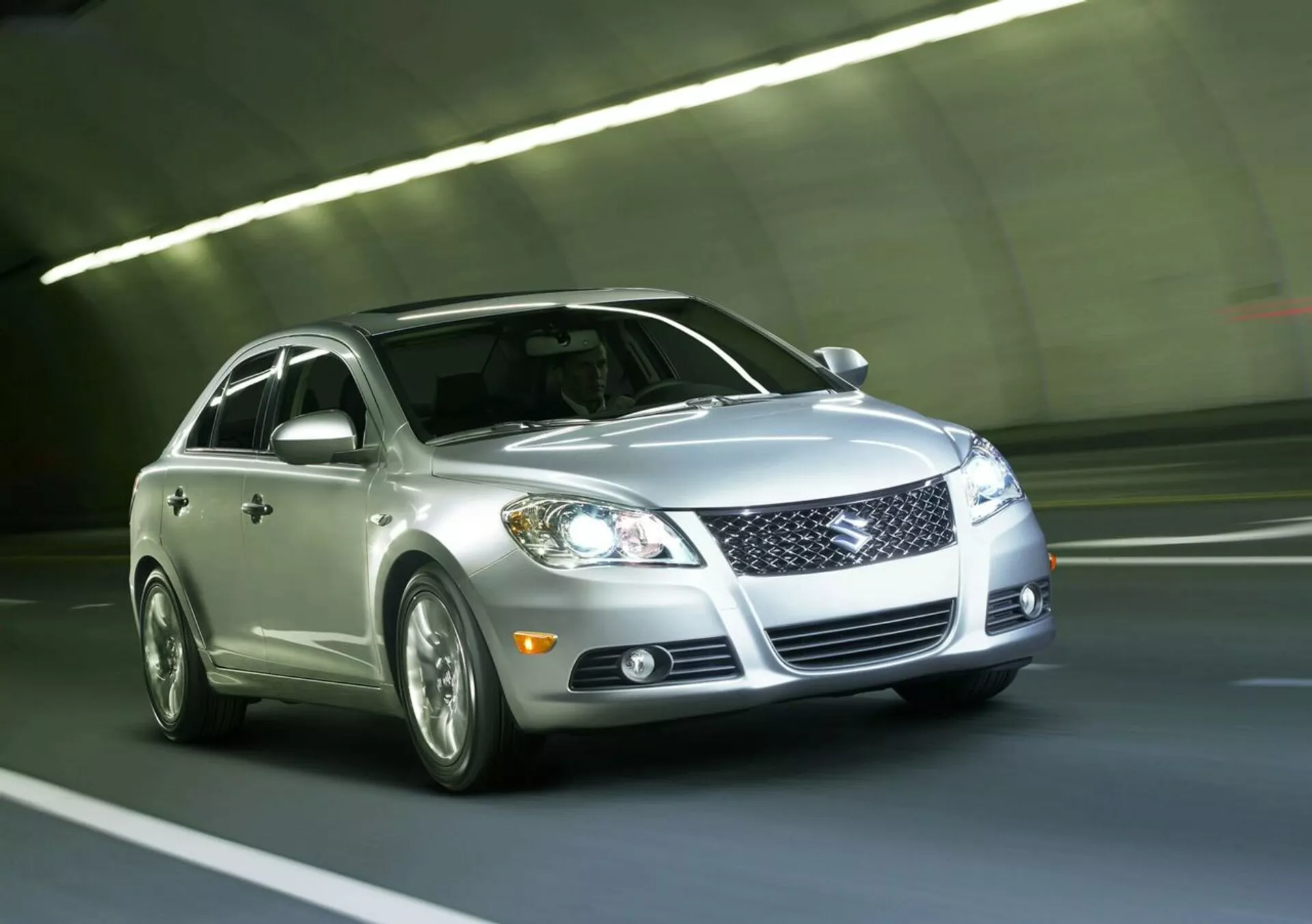 Stříbrný sedan Suzuki Kizashi 2010 projíždějící tunelem, zobrazující přední mřížku, LED světlomety, mlhová světla, slitinová kola a odrazivý lak, pohled z nízkého předního tříčtvrtečního úhlu zdůrazňující dynamický design a technologii vozidla.