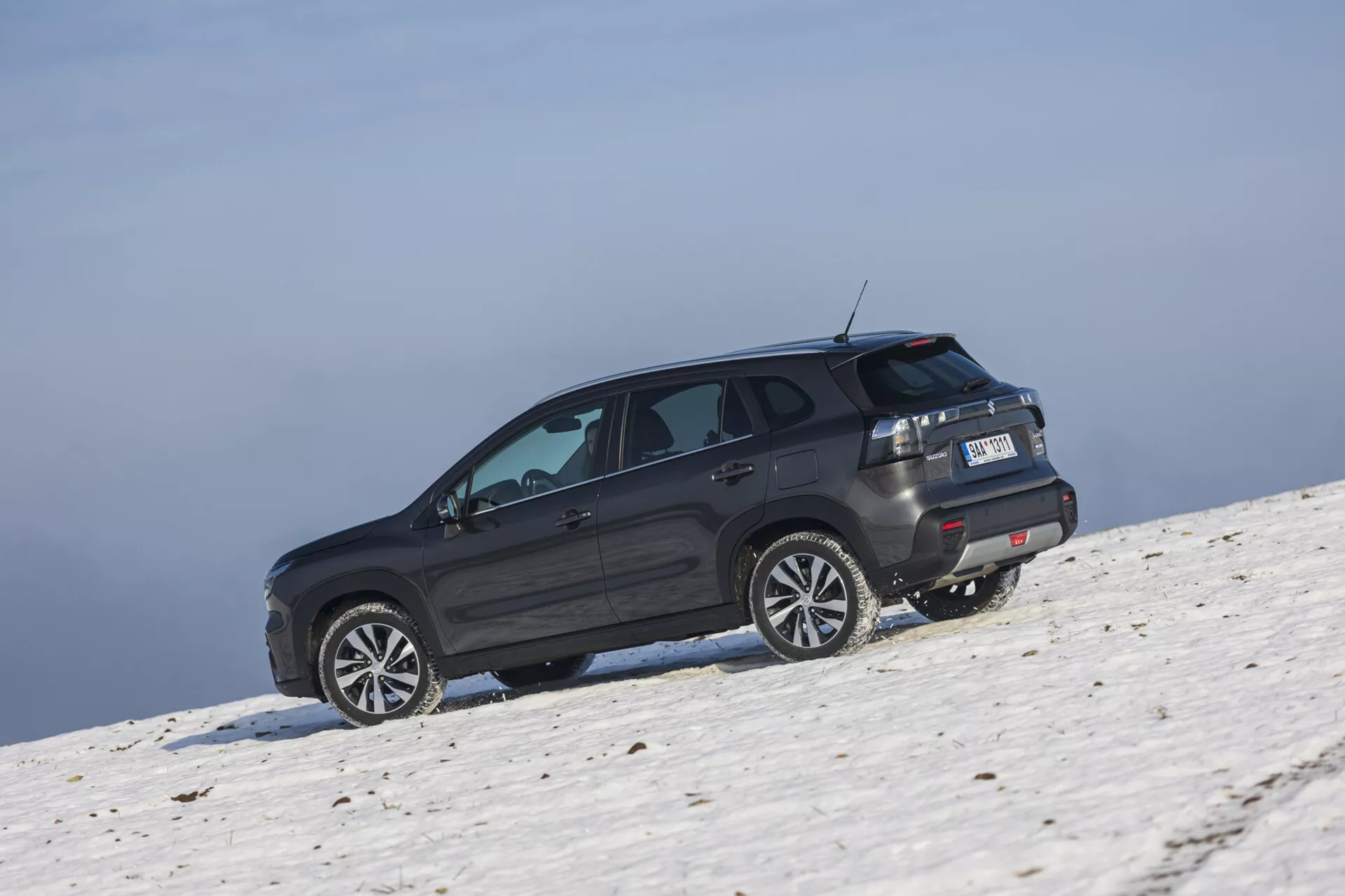 SUV Suzuki S-Cross 2022 v tmavém odstínu umístěné na zasněženém svahu zachycené z šikmého zadního úhlu, který zdůrazňuje jeho boční a zadní profil, velká slitinová kola, tónovaná okna s odmrazovačem, LED zadní světla, střešní spoiler a potenciální off-road schopnosti v podmínkách jasné oblohy.