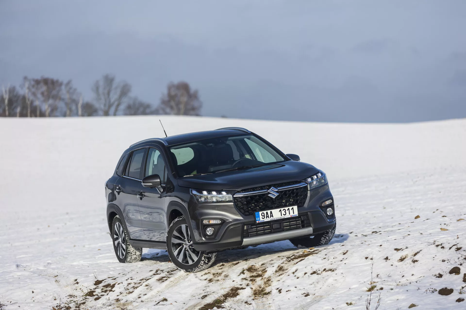 Tmavě zbarvené kompaktní SUV Suzuki S-Cross ročník 2022 zaparkované ve sněžné krajině, zobrazující pohled z předu a ze strany pod úhlem 45 stupňů, s elegantní mřížkou s kovovým povrchem, protáhlými LED světlomety, slitinovými koly s vícepaprskovým designem a leštěným chromovým rámečkem oken naznačujícím luxusní výbavu a potenciální schopnost terénní jízdy