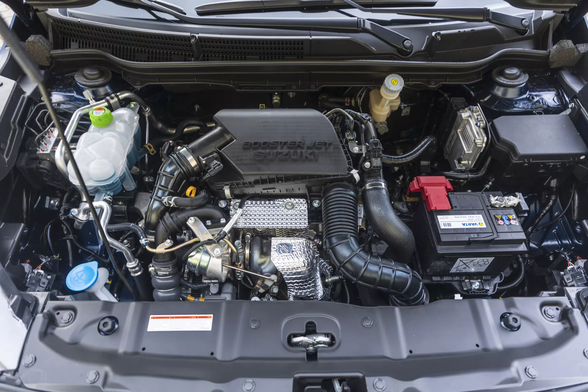 Pohled zepředu na motorový prostor Suzuki S-Cross 2022 s viditelnými součástmi jako jsou blok motoru, hlava válce, sací potrubí a technologickými prvky včetně turbodmychadla a proměnlivého časování ventilů, spolu se značením Suzuki na krytu motoru, v bezvadném stavu.