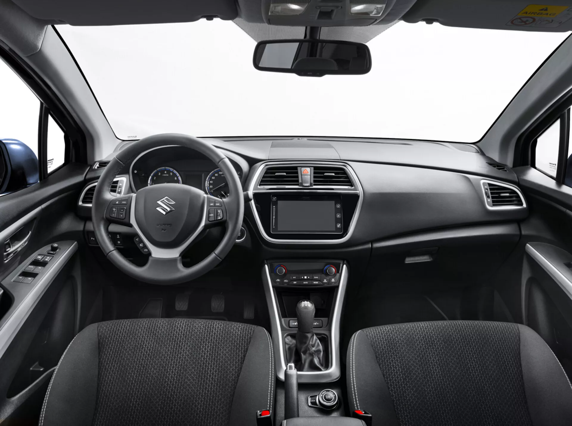 Interiér Suzuki S-Cross 2022 z pohledu zadního sedadla ukazující palubní desku, volant s ovládacími prvky, digitální přístrojový panel, systém infotainmentu, ovládání klimatizace, páku automatické převodovky a detailně propracovaná přední sedadla s texturou látky